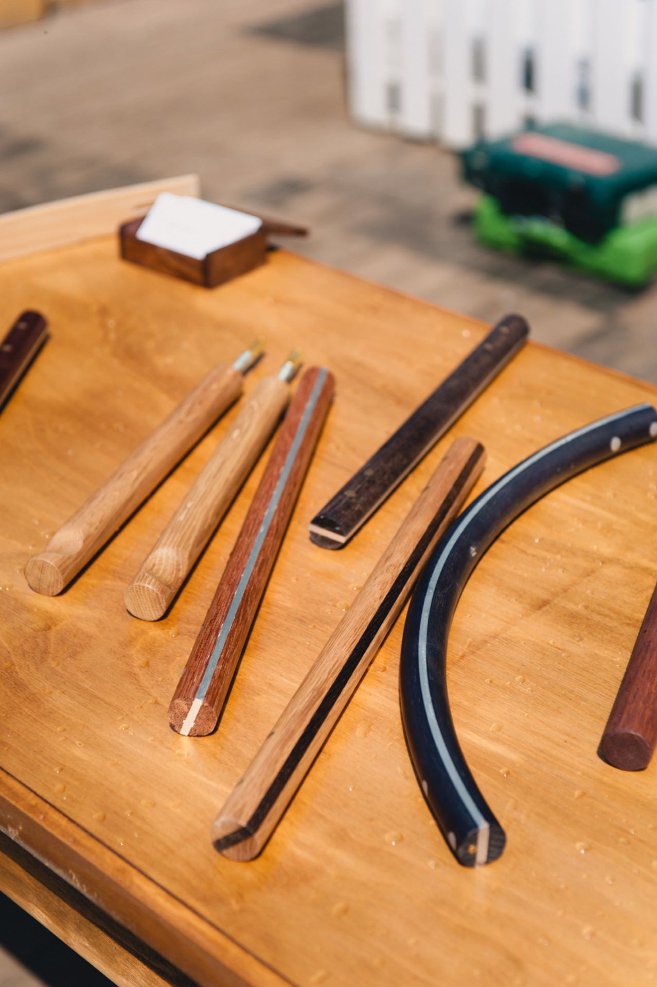 A wooden table displaying various woodworking tools and materials, including several wooden handles, a curved metal piece and neatly arranged wooden slats. A white fence and a green toolbox can be seen in the background of this photo scene from the fair. 