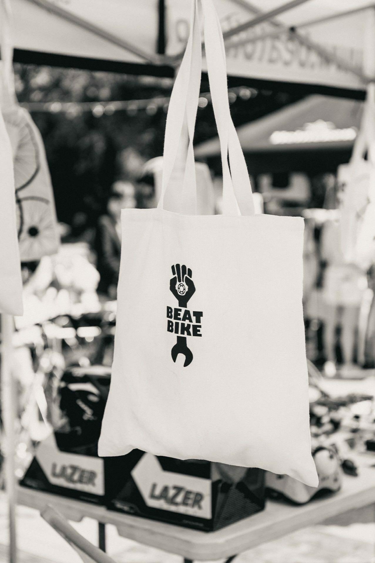 Black and white image showing a bag hanging on a stand. The bag features an illustration of an arm holding a key with the words "BEAT BIKE" printed on it. The blurred background suggests that it is part of a photo essay of a trade show, capturing the lively atmosphere of a fair or event setting.  