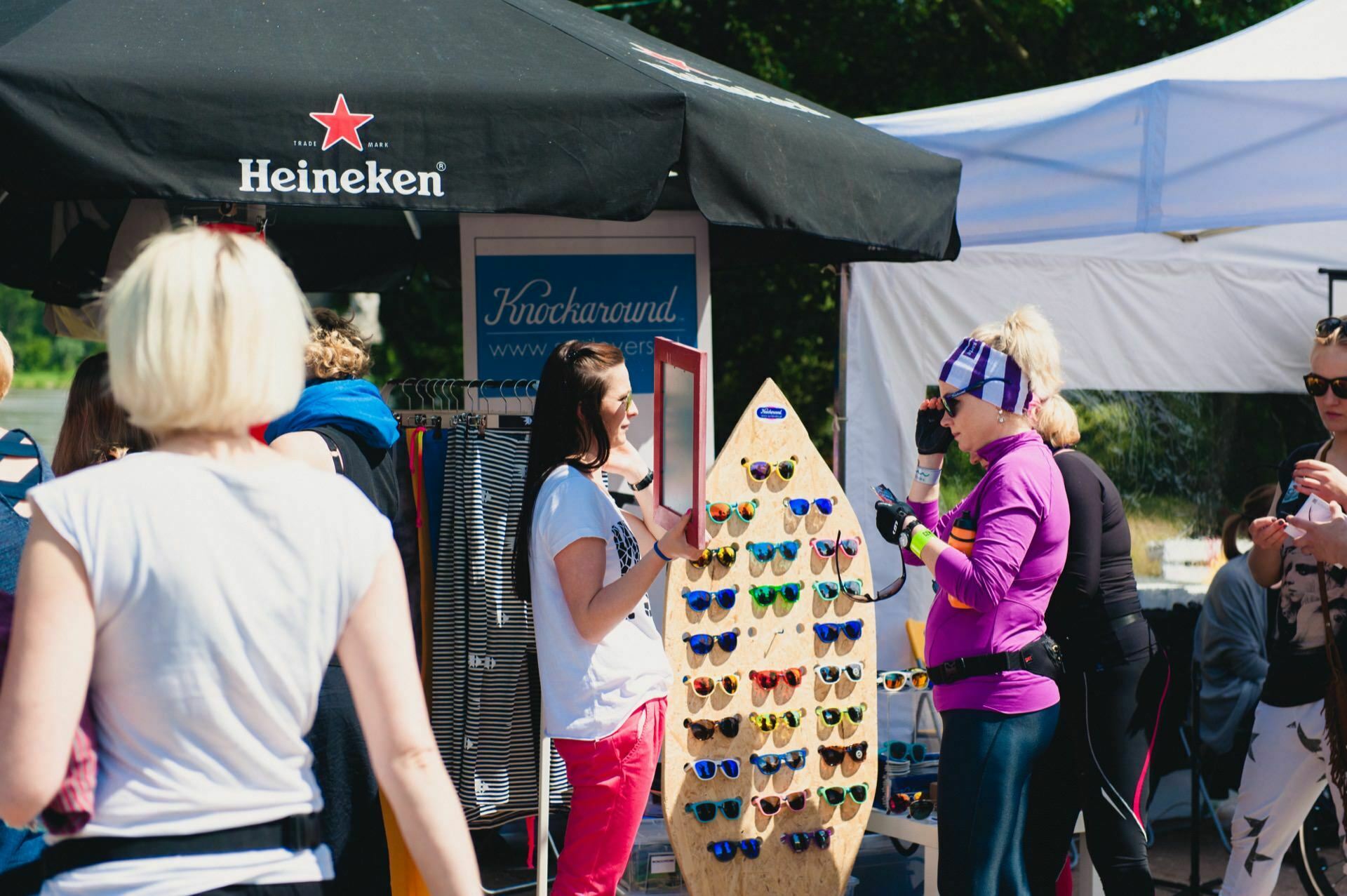 A group of people interact at an outdoor market with sunglasses set on a stand shaped like a surfboard, as captured in this photo essay from the market. Two women chat nearby while in the background a Heineken umbrella provides shade next to a blue sign. 