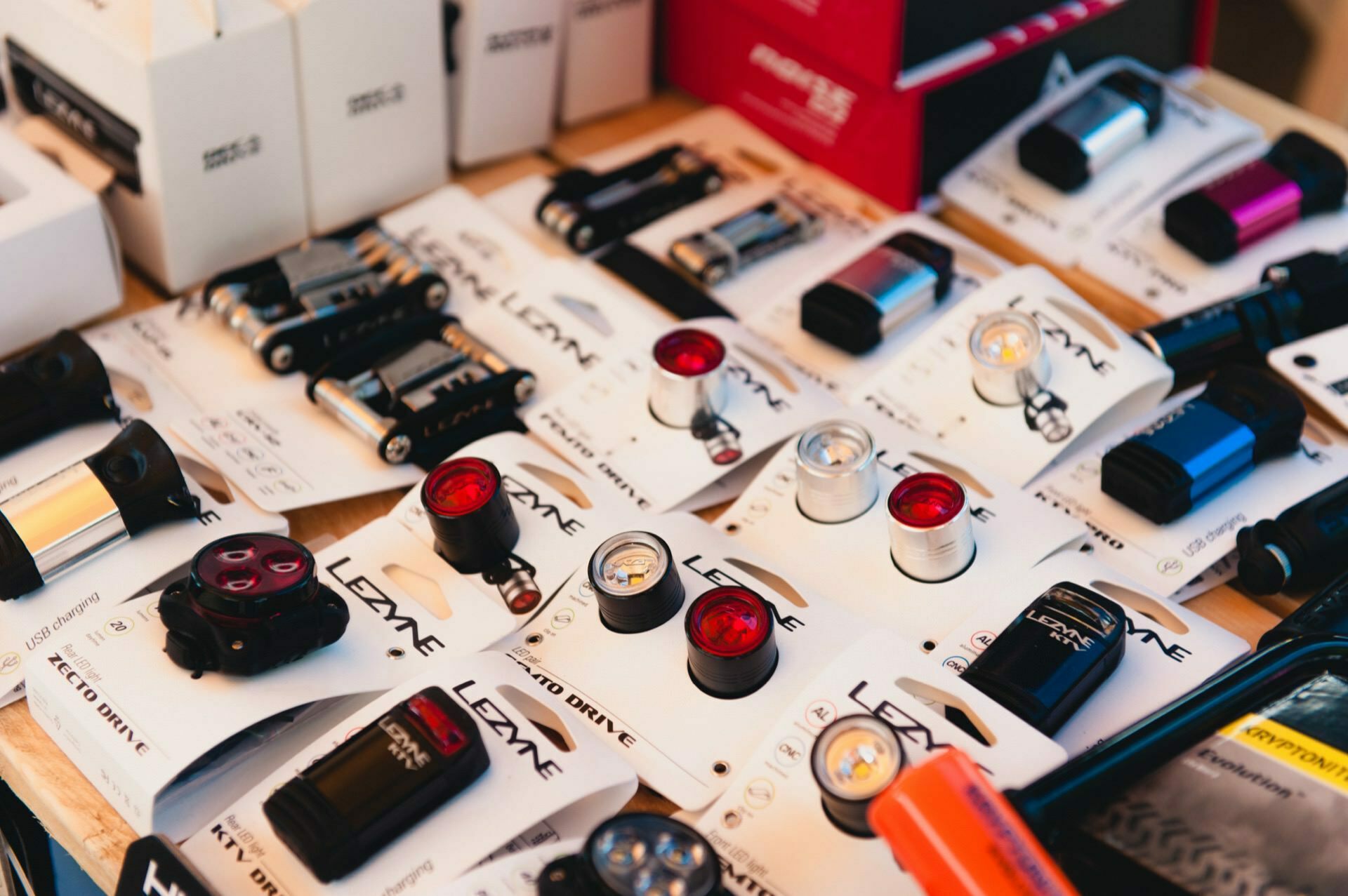 A display of various Lezyne bike lights and accessories in packs, captured by an event photographer. Products include hand lamps, headlamps and tail lamps in various colors and sizes, neatly arranged on a table. You can also see some of the boxes and packaging in the background of this photo report from the show.  