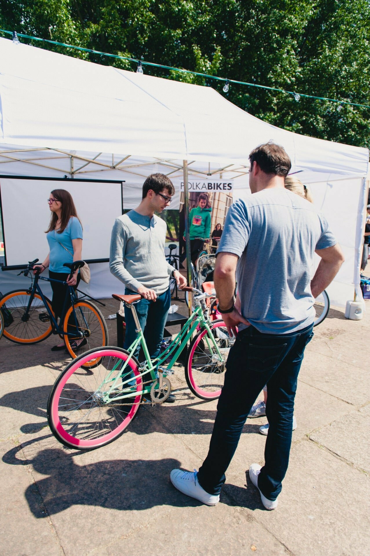 People gather around a bike shed outside, with a tent overhead. Two men are discussing, standing next to the colorful bicycles. A woman holding a bicycle stands nearby, and another woman can be seen in the background. Trees are visible, indicating the park's surroundings - the perfect scene for any event photographer to photograph the fair.   