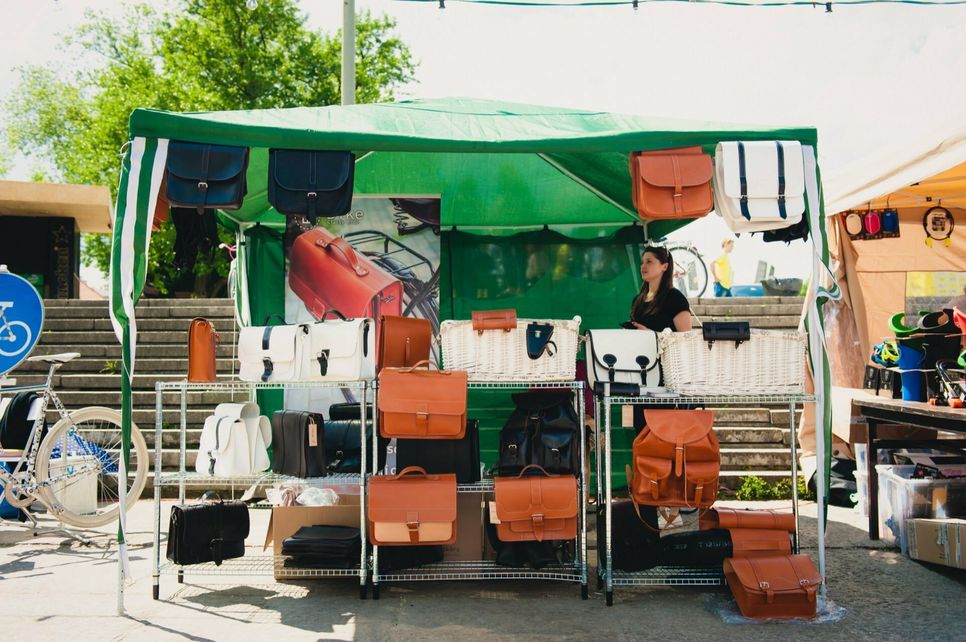 A woman stands at a green outdoor stall displaying a variety of leather bags. The bags in shades of brown, black and white are neatly arranged on shelves. Bicycles and trees can be seen in the background, suggesting a vibrant outdoor setting, beautifully captured by the event photographer.  