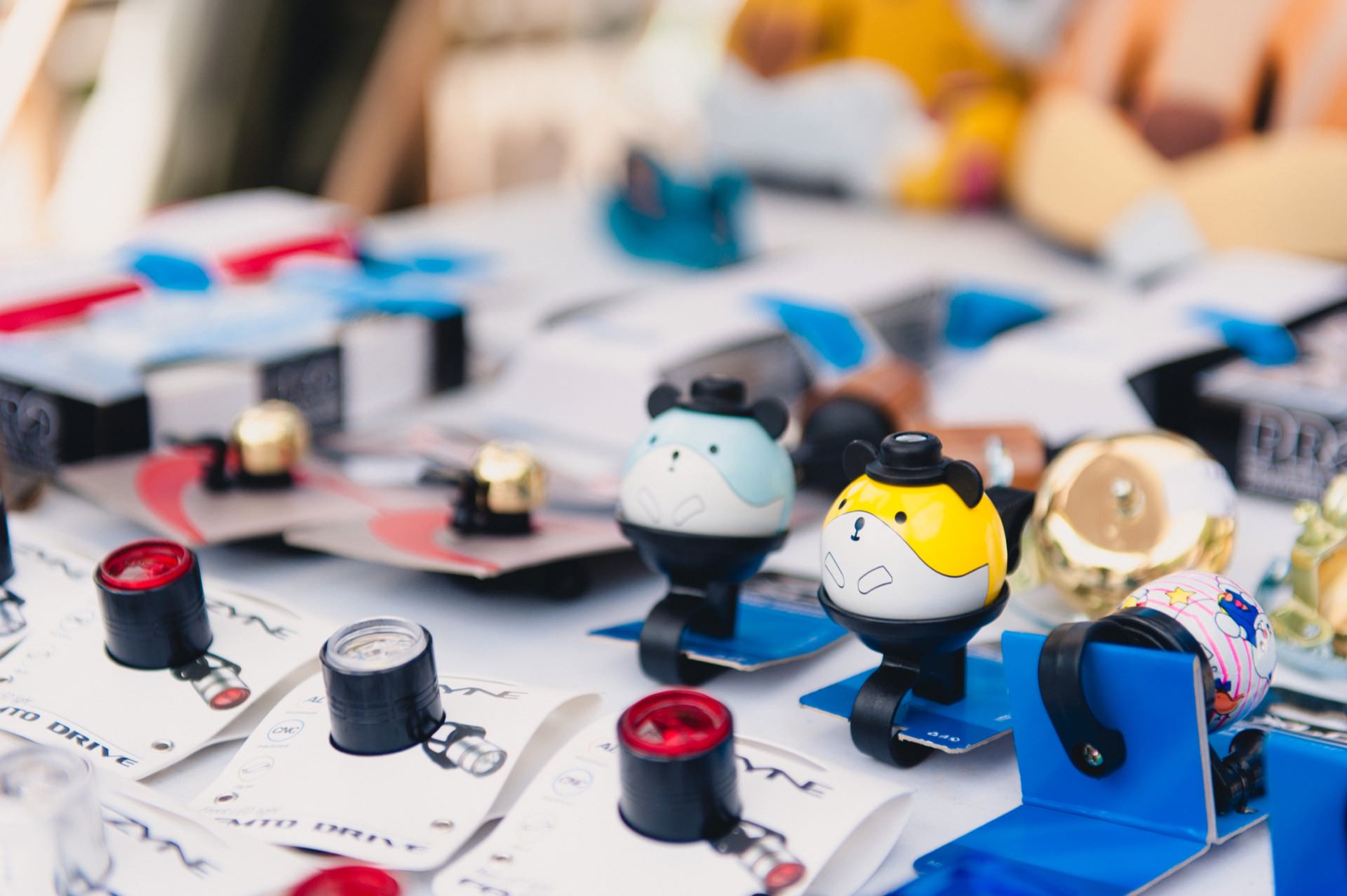 A display of colorful and fun bicycle bells, including designs resembling cartoon characters, is set up on a table. Other bicycle accessories and packaging can also be seen in the background, suggesting the setting of a market or store, perfect for a vibrant photo essay of the fair. 