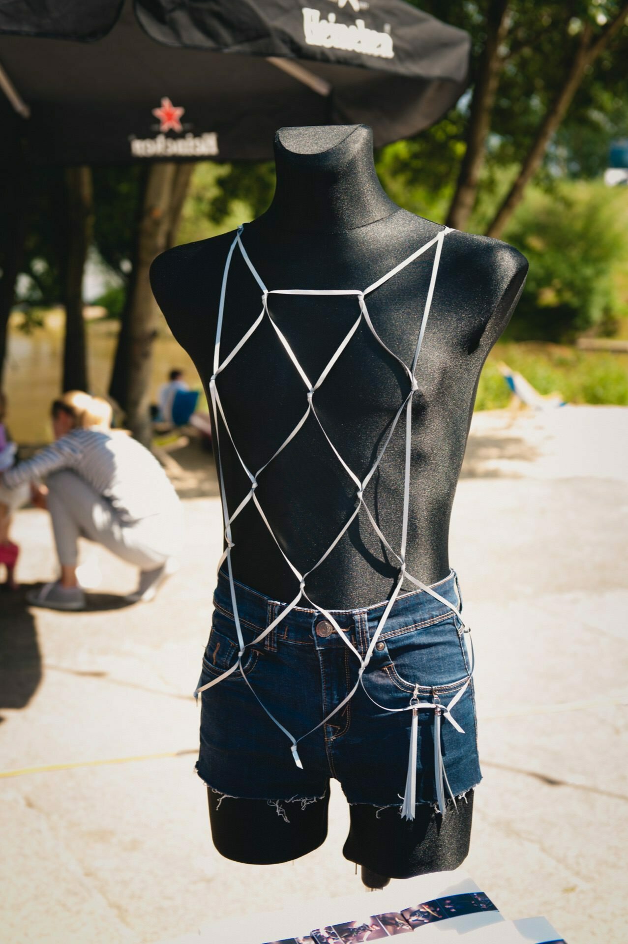 The black mannequin is dressed in a white mesh top and denim shorts. The mannequin is displayed outside with an umbrella and greenery in the background. People can be seen in the distance, but they are out of focus, capturing the perfect moment for a photo fair.  