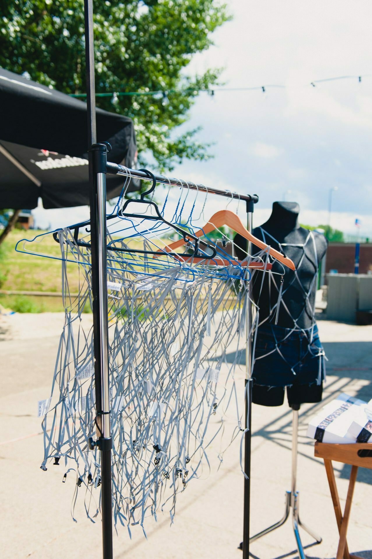 The exterior configuration depicts a black mannequin dressed in a net-like outfit and a clothes rack filled with several hangers draped in similar net material. A table and canopy are partially visible in the background, with greenery and blue skies in the distance - capturing the essence of a captivating photo fair. 