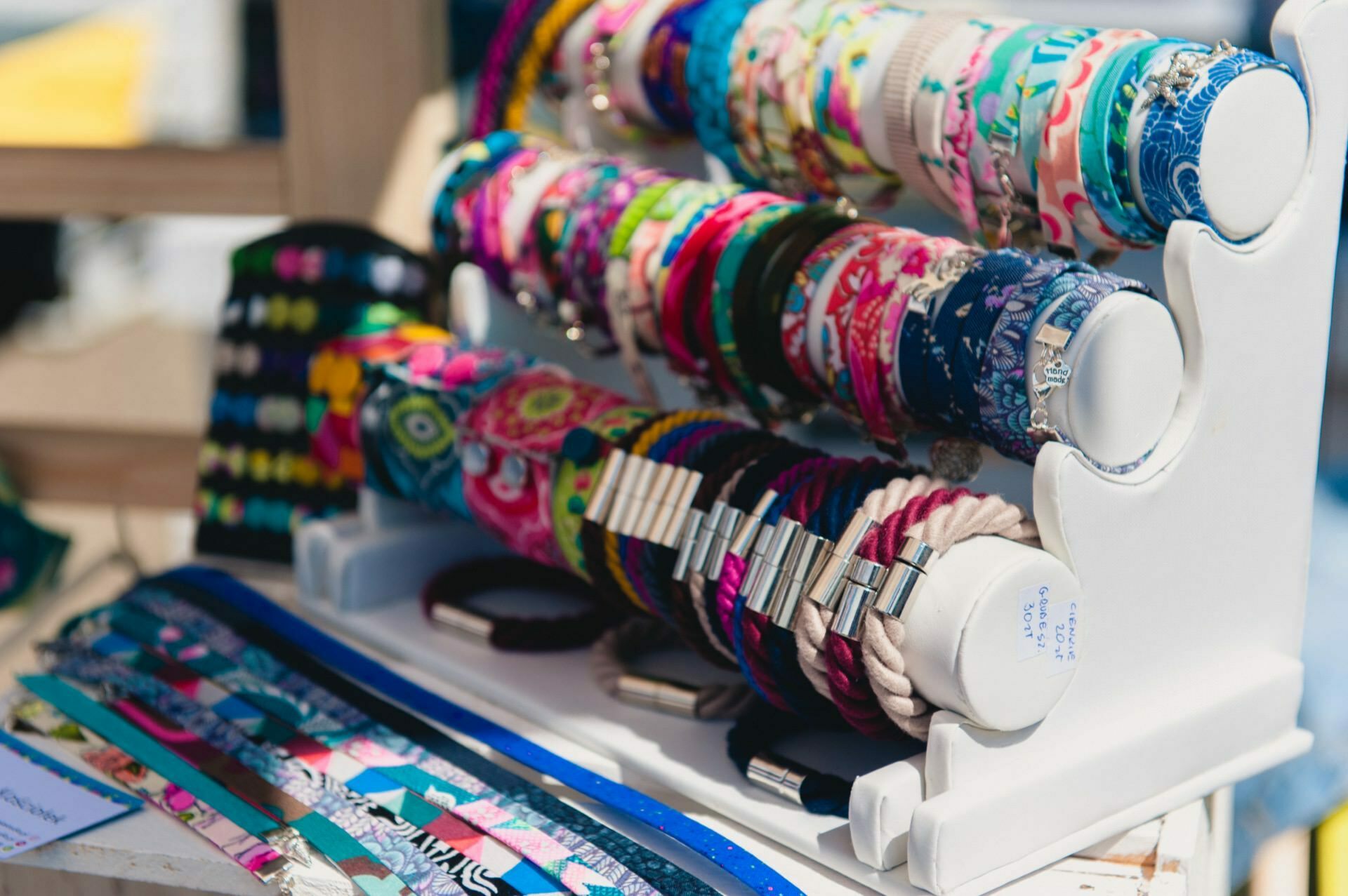 An exhibition of colorful patterned bracelets and headbands arranged on a white stand catches the eye. The vibrant designs shine in the sunny surroundings of the marketplace, perfect for photo opportunities at the fair. In the display case you'll find both thicker headbands and sleeker, elegant options, the perfect material for any event photographer.  