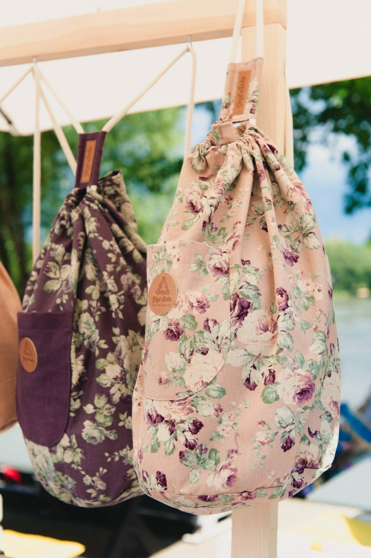 Two floral string bags hang side by side on a wooden stand. The bag on the left has a dark purple background with pink and white flowers, while the one on the right has a light pink background with pink and white flowers. In the background you can see a picturesque outdoor setting, perfect for a photo fair.  