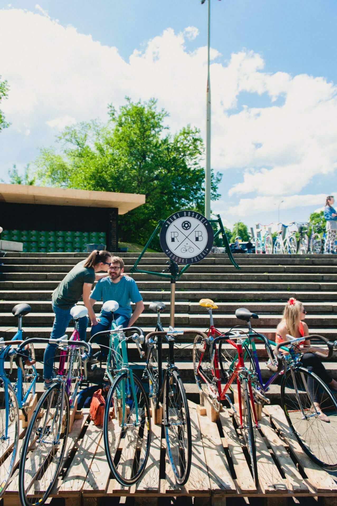 An outdoor scene captured by an event photographer shows several bicycles parked in front of a stairwell. Two people are interacting near the bikes, a child is sitting on the stairs to the right. In the foreground is a circular sign with letters, with trees and clear sky in the background.  