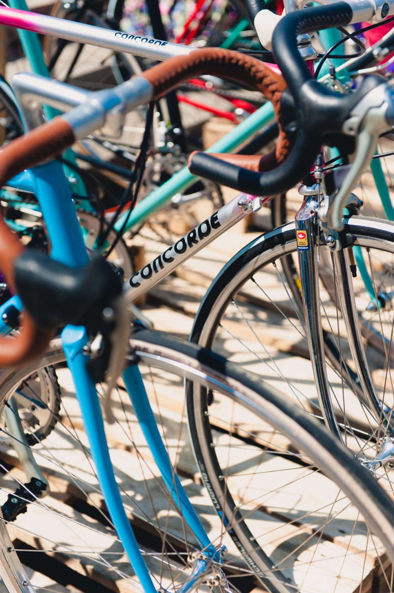 Close-up of several parked bikes, focusing on one blue bike with brown tape on the handlebars and "Concorde" branding on the frame. Other brightly colored bicycles can be seen in the background with a vibrant photo of the fair. The sun casts shadows, indicating the setting outside.  