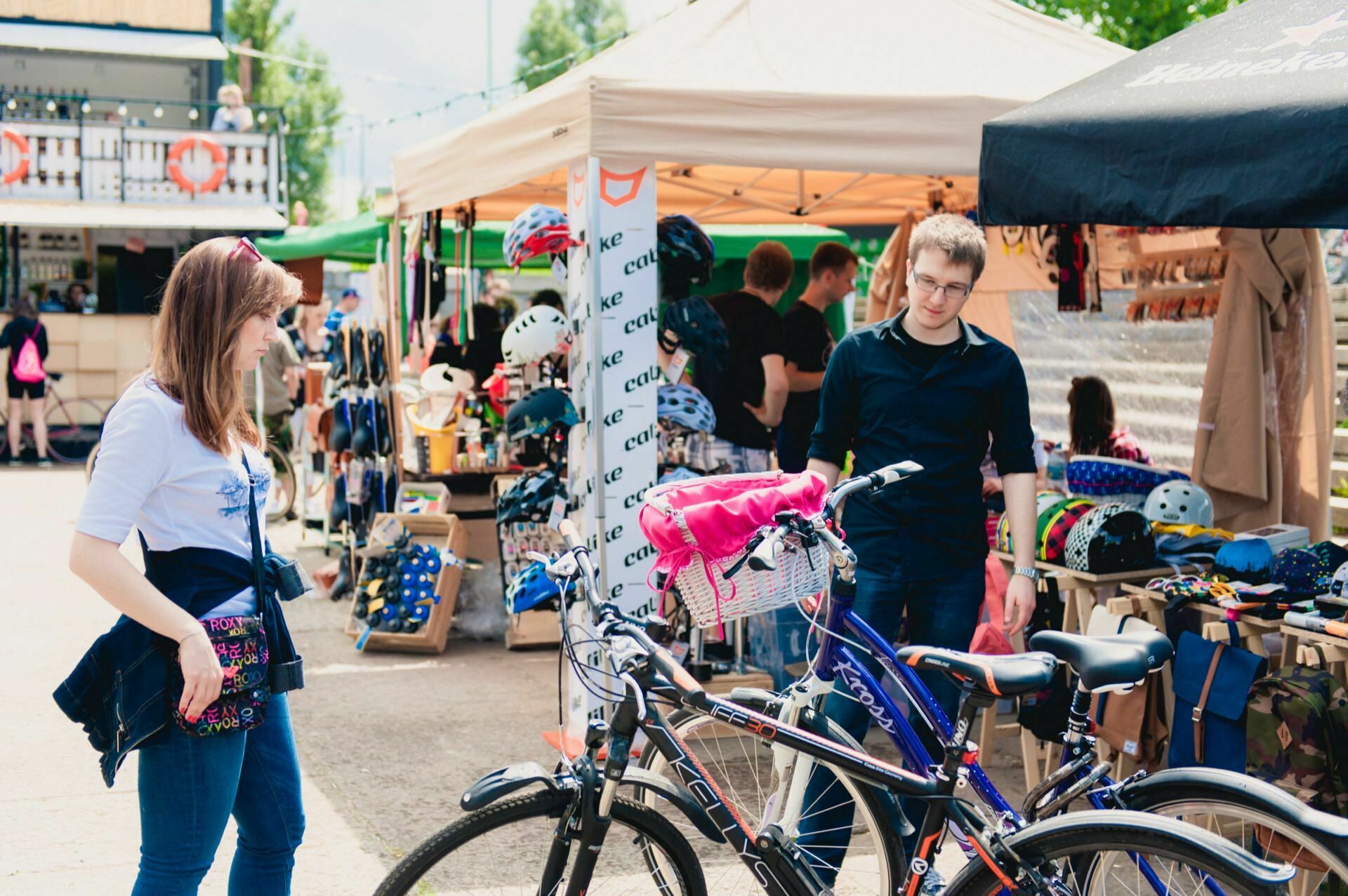 Two people on an outdoor stand are looking at a bicycle. The woman on the left is wearing a white shirt and blue jeans, while the man on the right is wearing glasses and a dark shirt. Various items for sale are displayed under the tents, including helmets and accessories. This photo could be part of a photo essay of the fair taken by an experienced event photographer.   