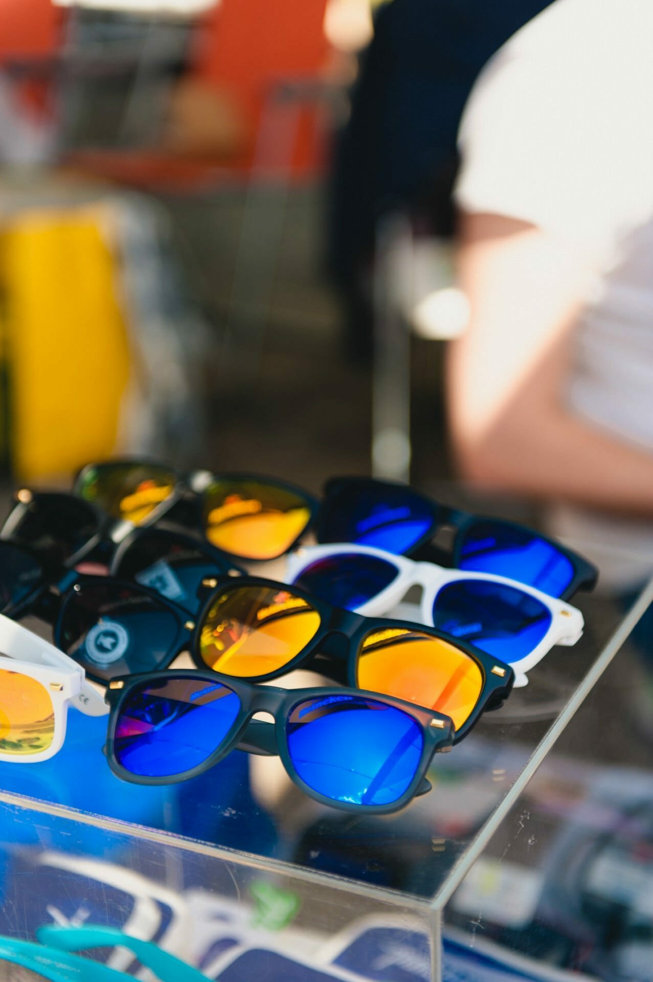 Presenting various sunglasses with reflective lenses in colors such as blue and orange. Some frames are black, while others are white. The glasses are placed on a transparent acrylic stand with a fuzzy background indicating an open-air setting - perfect for a trade show photo essay.  