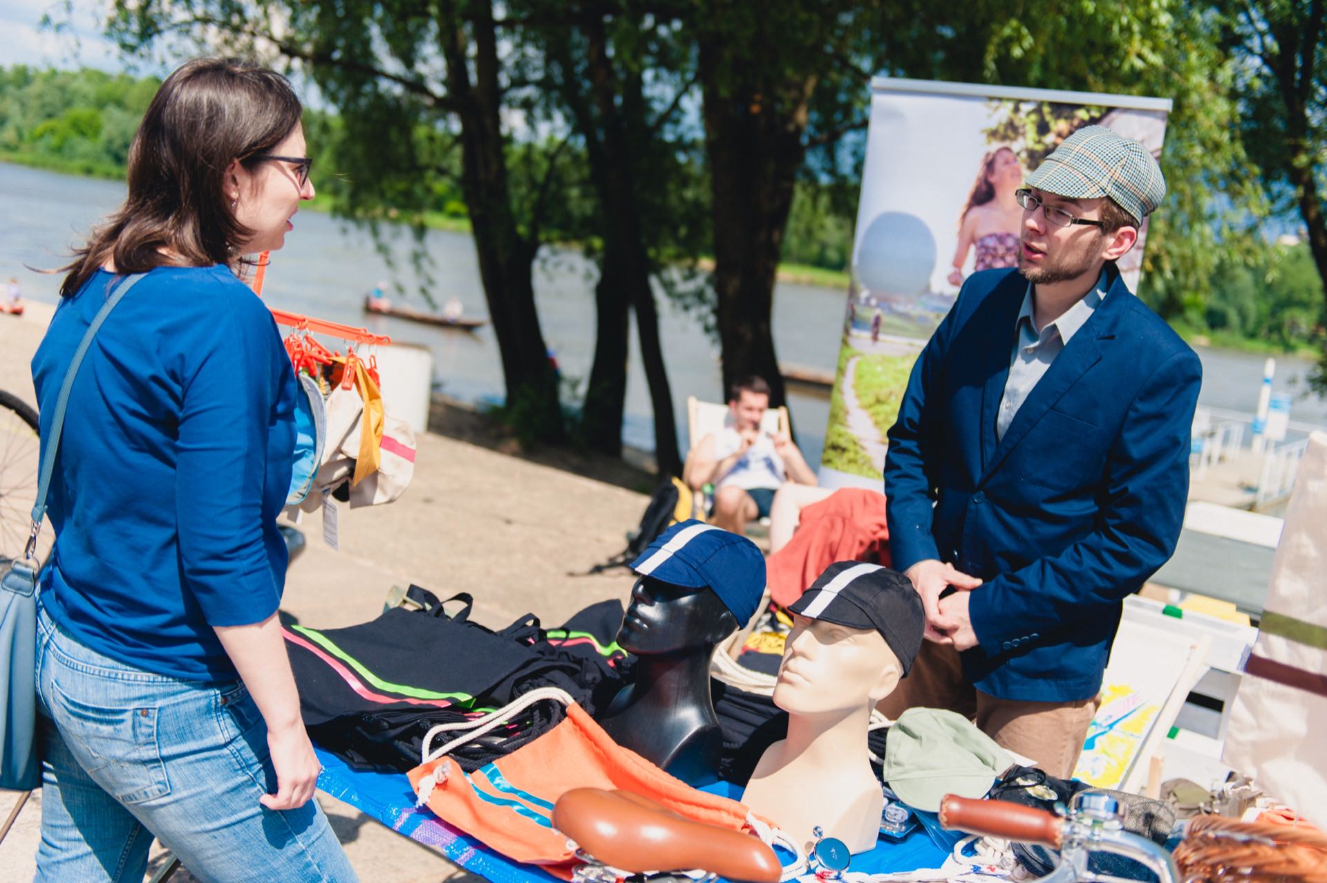 A woman wearing glasses talks to a man wearing a hat at a stall by the river. The stall displays a variety of items, including hats, bags and bike seats. In the background you can see the trees and landscape of the river banks, beautifully captured in this photo essay of the fair. A person lies on a chair nearby.   