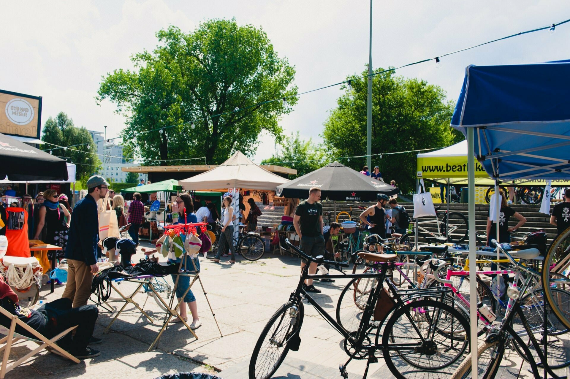 The bustling outdoor market, captured in a lively photo essay, shows bicycles, tents and people mingling. Vendors display items under canopy tents, while cyclists gather near parked bicycles. Trees provide a lush backdrop, and sunlight brightens the scene - a perfect photo essay of the fair.  