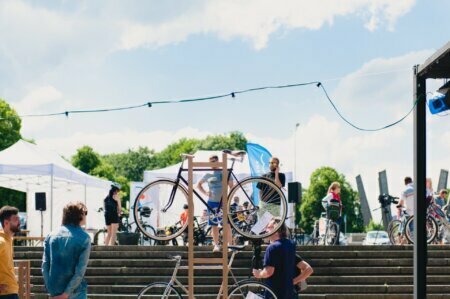 Impreza plenerowa z publicznością skupioną wokół ekspozycji rowerów, w tym jednego zamontowanego na drewnianym stojaku. Na scenie w tle ktoś mówi do mikrofonu. Nad głowami wiszą świąteczne girlandy świetlne, które oddają ożywioną atmosferę, idealną dla każdej fotorelacji z targów. Zielone drzewa i słoneczne niebo w tle.