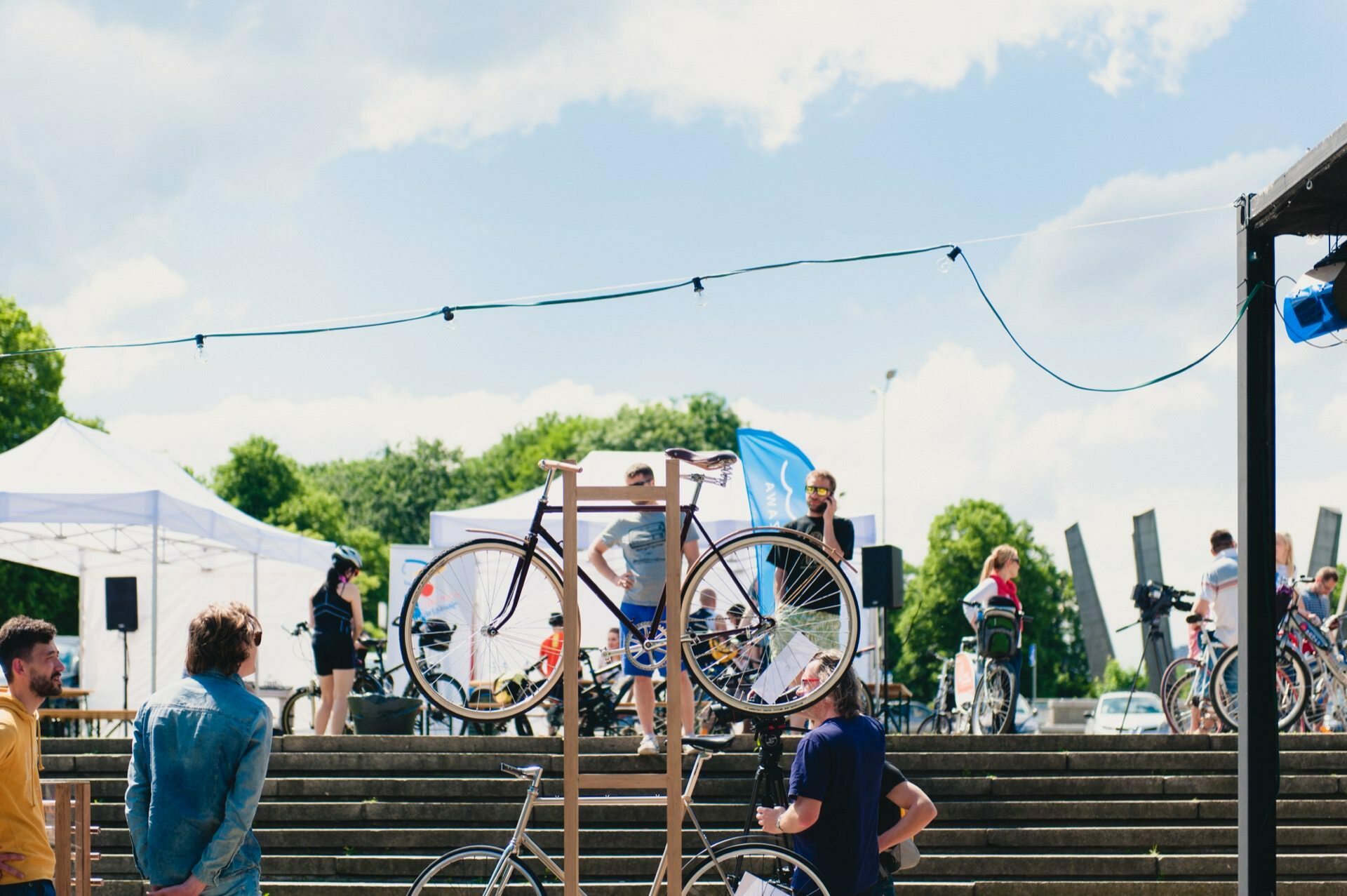 An outdoor event with the audience gathered around a display of bicycles, including one mounted on a wooden stand. On a stage in the background, someone speaks into a microphone. Christmas light garlands hang overhead, capturing the lively atmosphere that is perfect for any photo fair. Green trees and sunny skies in the background.   