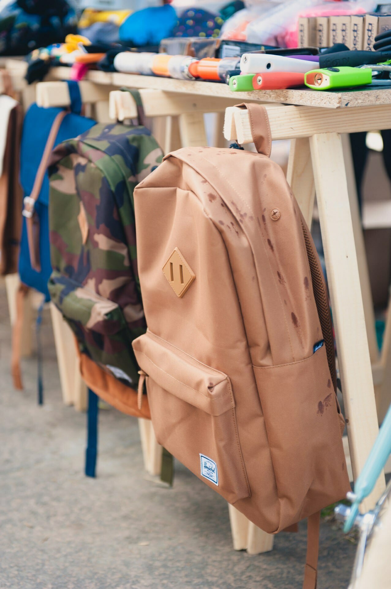 An exhibition of backpacks hanging on a wooden stand at an outdoor market, captured as part of a photo report of the fair. In the foreground you can clearly see a brown backpack with what looks like water stains, and next to it you can see a backpack in a camouflage pattern. There are various items on the table above.  