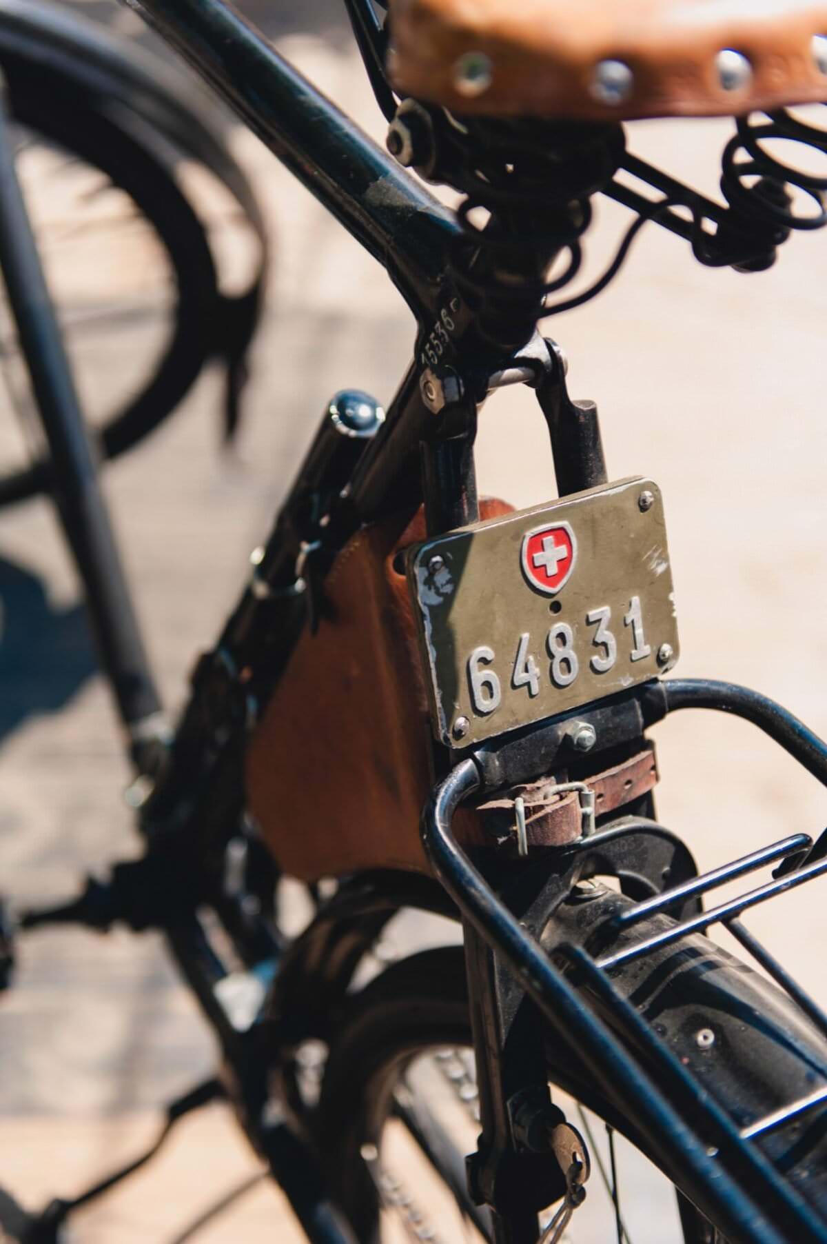 Zbliżenie na czarny zabytkowy rower ze szwajcarską tablicą rejestracyjną z napisem „64831” i emblematem szwajcarskiej flagi. Siedzisko ma skórzaną wyściółkę, a drewniana skrzynia przednia jest częściowo widoczna. To idealny obiekt dla każdego fotografa eventowego, który uchwyci misterne szczegóły na rozmytym tle poprawiającym ostrość.
