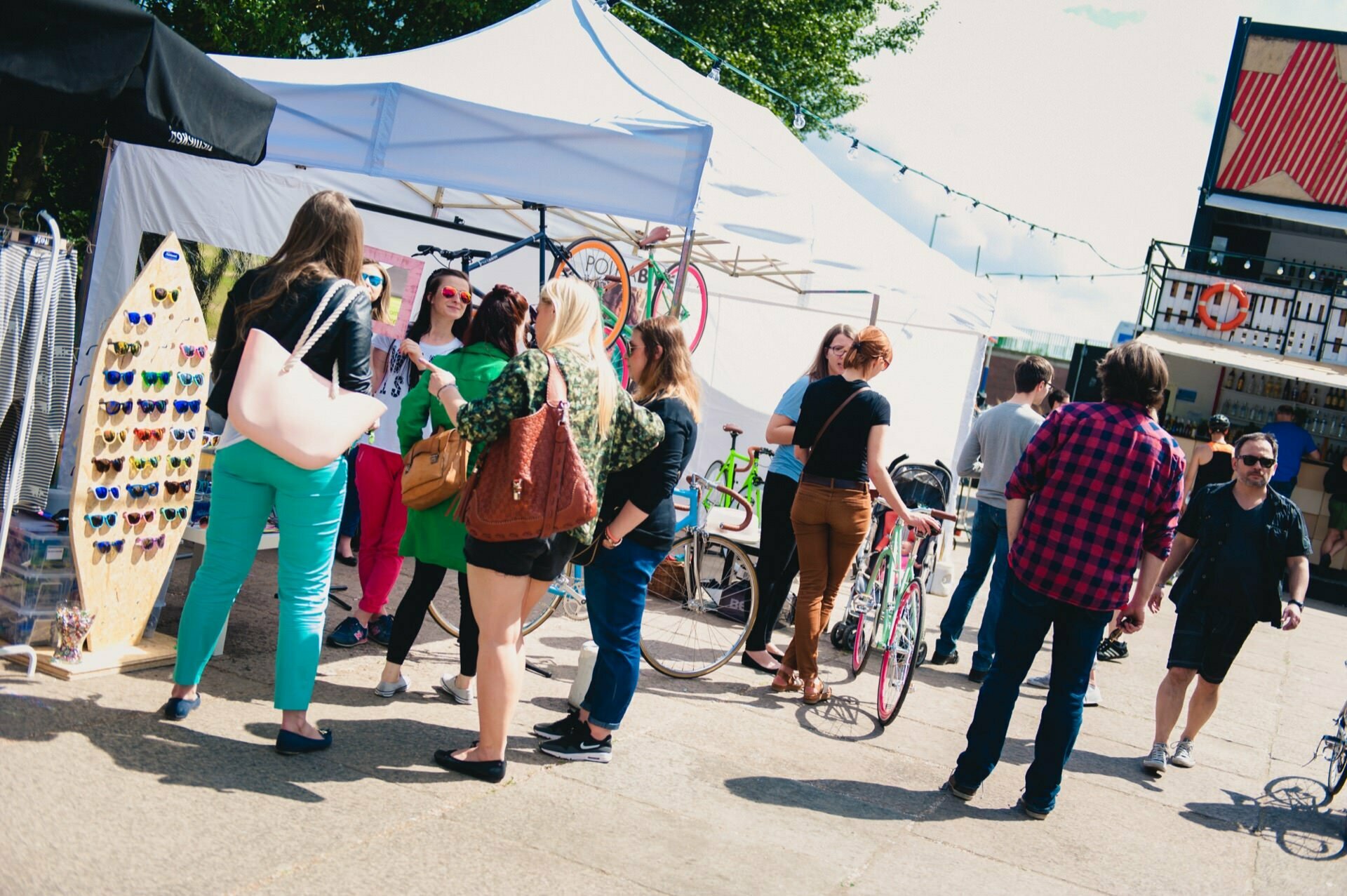 A group of people are standing and talking in an outdoor market. There are white tents set up with various items for sale, including a rack of sunglasses. Bicycles can be seen in the background, and a few more people are walking nearby. This scene is perfect for a photo report of the fair by an event photographer.   