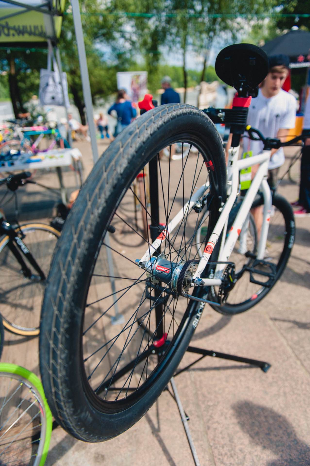 At an outdoor event, a man looks at a white bicycle mounted on a repair rack and captures the moments for a photo essay of the fair. The bike's rear wheel is visible in the foreground. Other bikes and people can be seen in the background under the tents, while trees provide shade.  