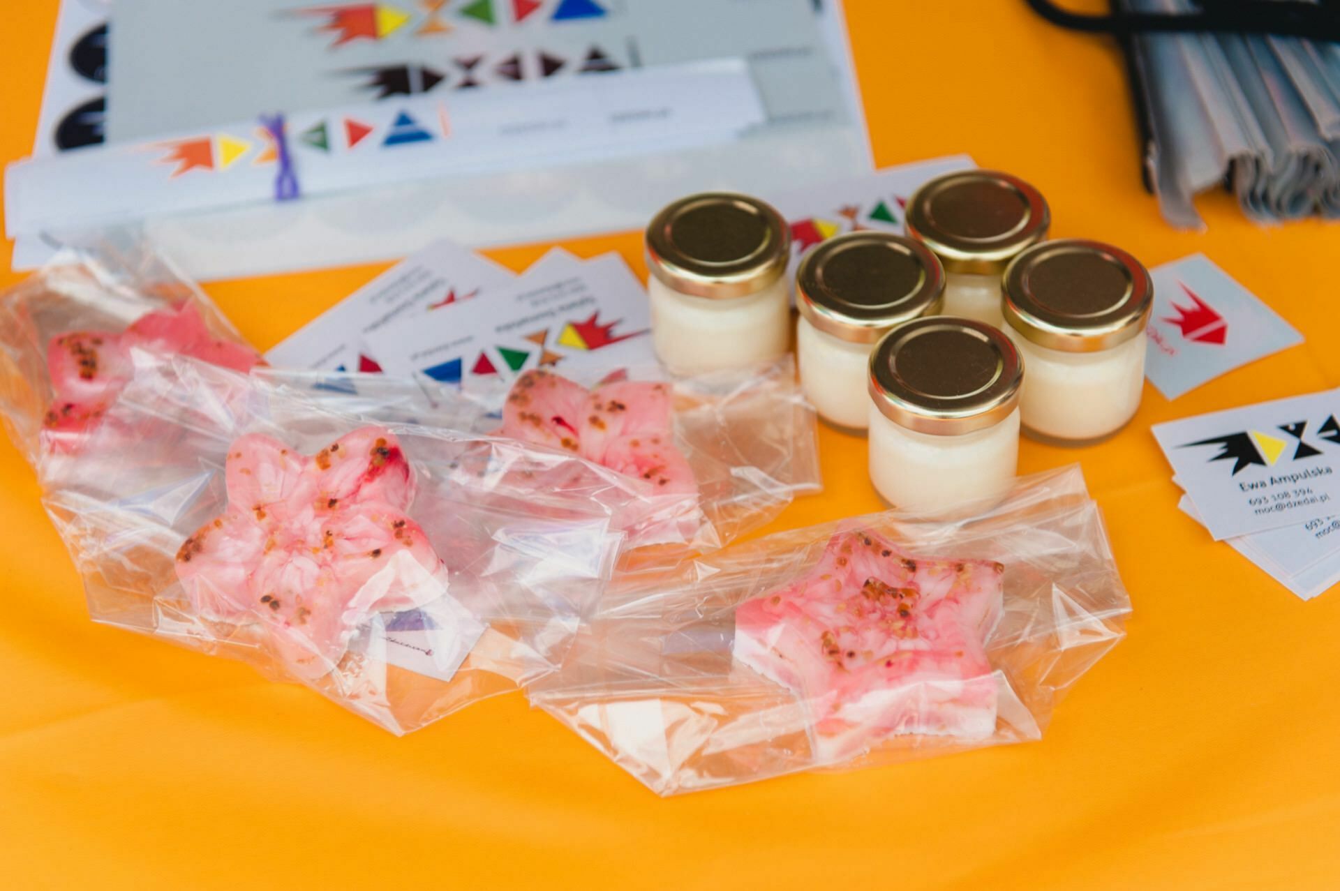 The display on the orange table contains small glass jars with gold lids, sealed packages of pink flower-shaped soaps and business cards with colorful geometric logos. Some of the documents and other materials are partially visible in the background, capturing the beautiful photo-op of the fair. 