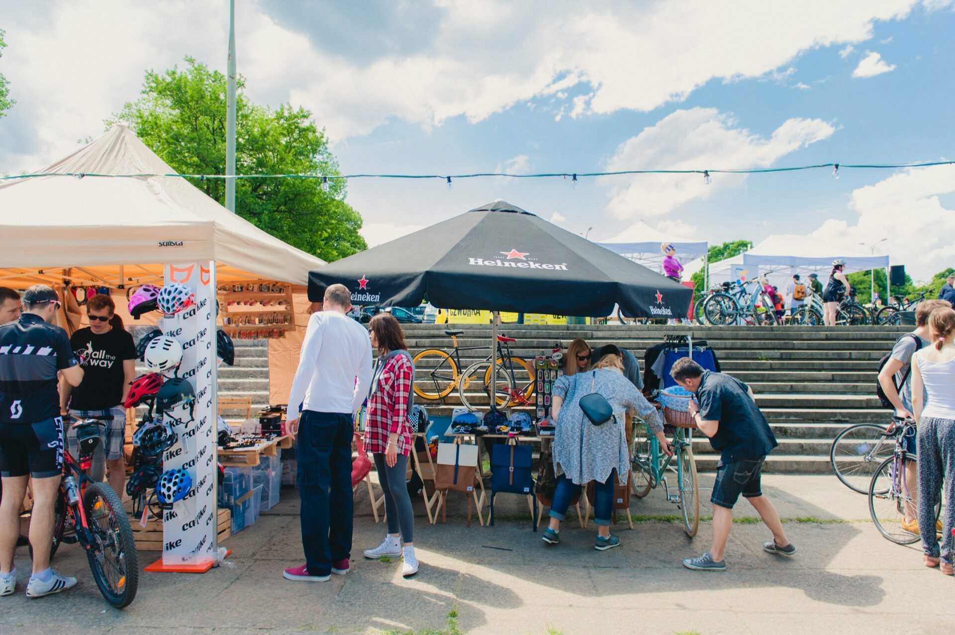 A bustling outdoor market scene where people are browsing various booths. One booth showcases bicycle helmets and accessories, while another, sheltered by a Heineken umbrella, displays bicycles. In the background, under a partly cloudy sky, you can see a staircase and a green tree - the perfect moment for a photo report of the fair.  