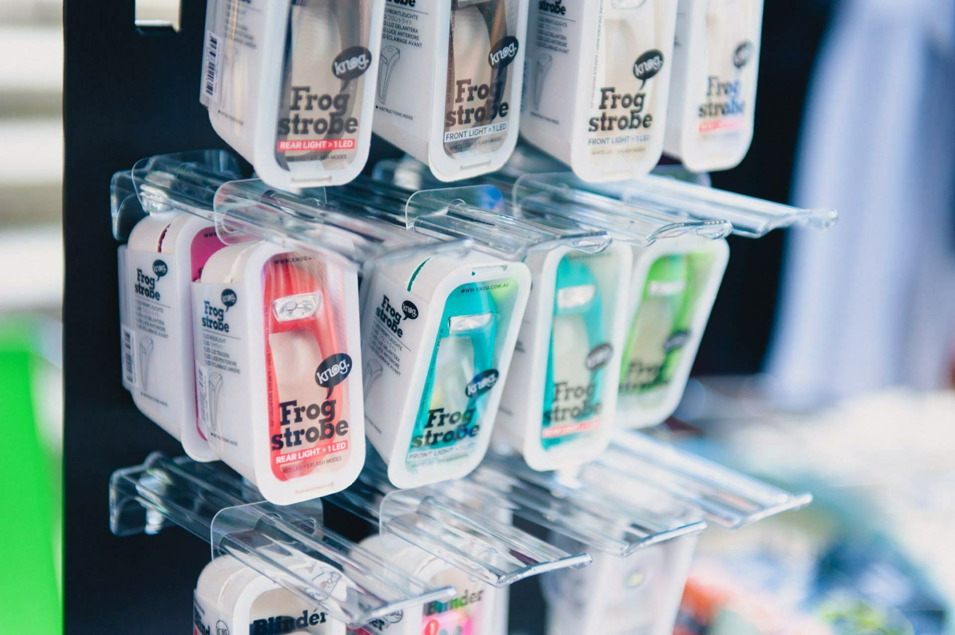 The display stand features a number of packages of bicycle lights with the words "Frog Strobe." The packages, made by an event photographer, come in a variety of colors, including red, blue, green and clear. Each package contains a compact frog-shaped bicycle light, perfectly framed like a photo fair.  