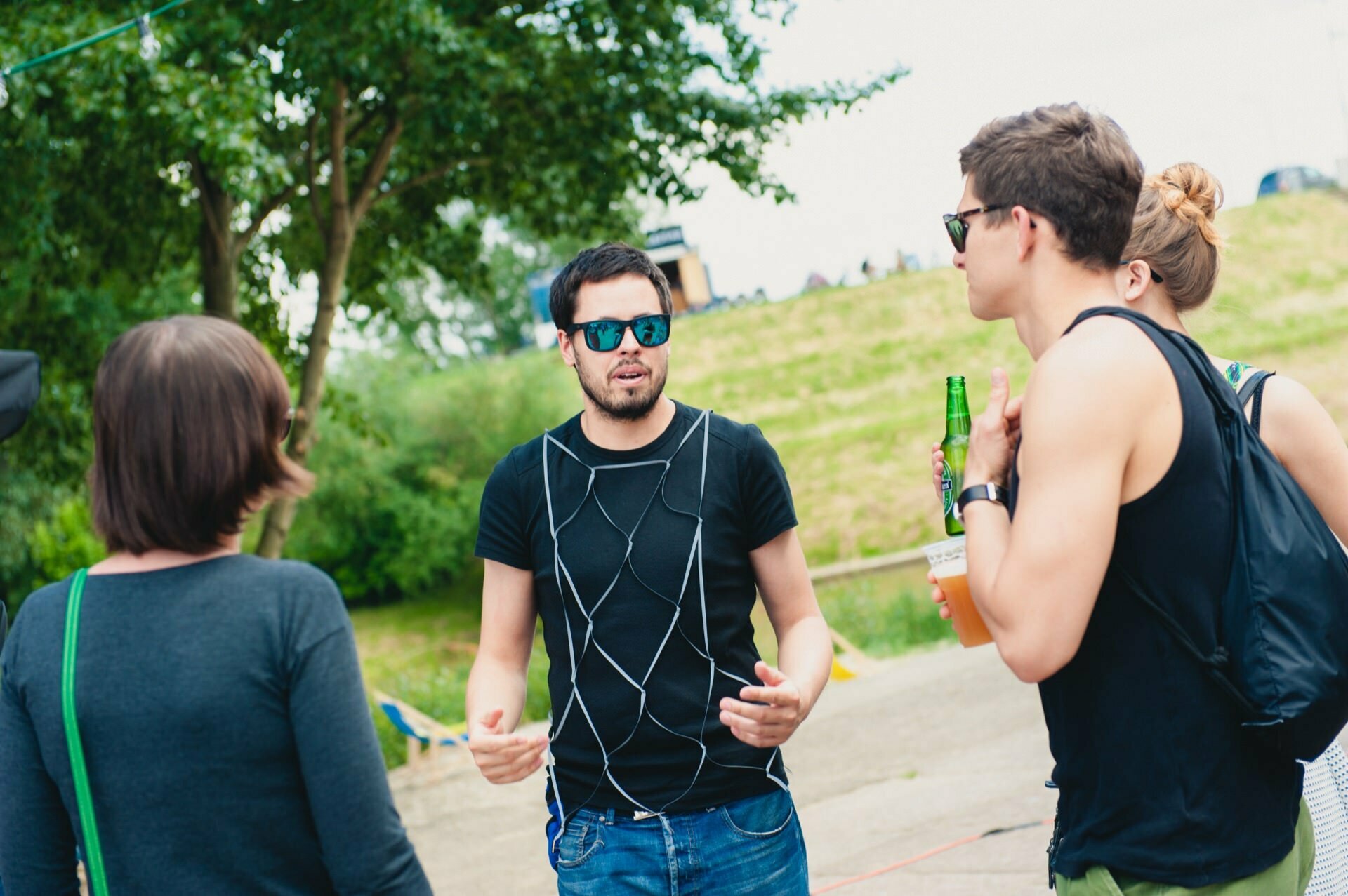 A group of people are standing outside talking. The man in the middle is wearing a black shirt and sunglasses and gesturing with his hands. Other people are holding drinks, and a grassy area with trees can be seen in the background. It appears to be a casual gathering captured by an event photographer.   