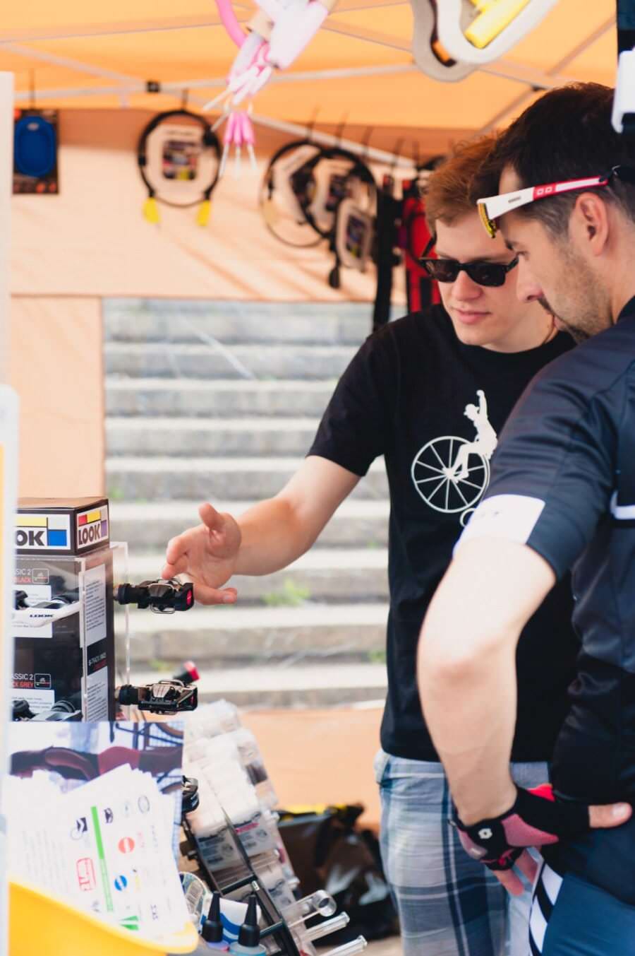 Dwóch mężczyzn stoi przy stoisku na świeżym powietrzu z akcesoriami rowerowymi. Jeden mężczyzna w czarnym t-shircie wyjaśnia produkt innemu mężczyźnie ubranemu w strój rowerowy. Na stoisku można zobaczyć różnorodne artykuły rowerowe i materiały promocyjne, które doskonale uchwyciła fotorelacja z targów. W tle widoczne są kroki.