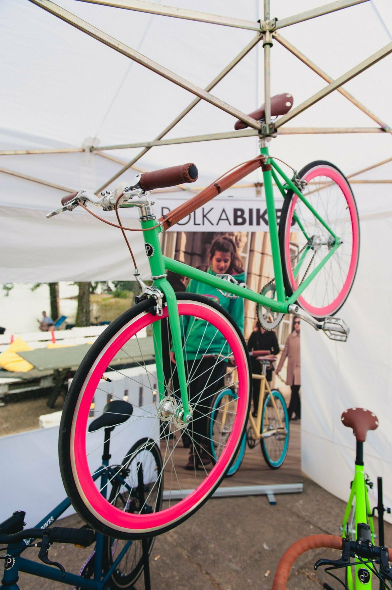 During the outdoor event, a green bicycle with pink tires is suspended under a white canopy. The scene is captured by an event photographer, with several other bikes and equipment in the background, and a "POLKABIKES" banner with an image of a person with a bicycle - perfect for a photo essay of the fair. 