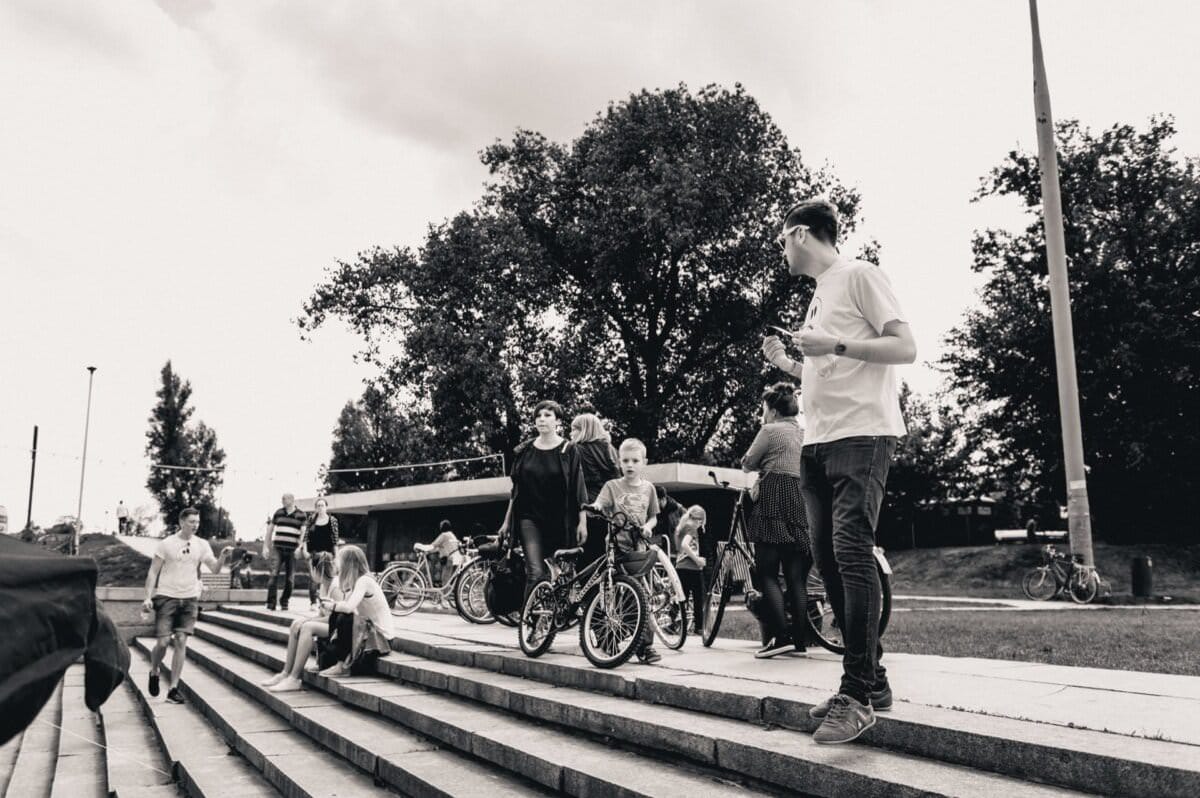 Grupa ludzi gromadzi się na zewnątrz na schodach w pobliżu rowerów. Kilkoro dzieci i dorosłych stoi, rozmawia i siedzi na schodach. W tle znajdują się duże drzewa i konstrukcja. Obraz, uchwycony przez wprawnego fotografa eventowego, jest czarno-biały.