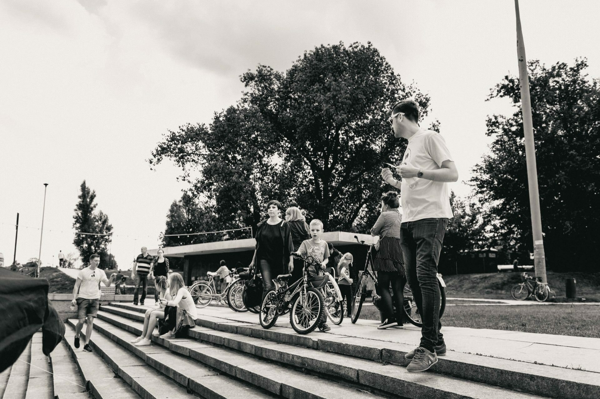 A group of people gather outside on the steps near the bicycles. Several children and adults are standing, talking and sitting on the steps. In the background are large trees and a structure. The image, captured by a skilled event photographer, is in black and white.   