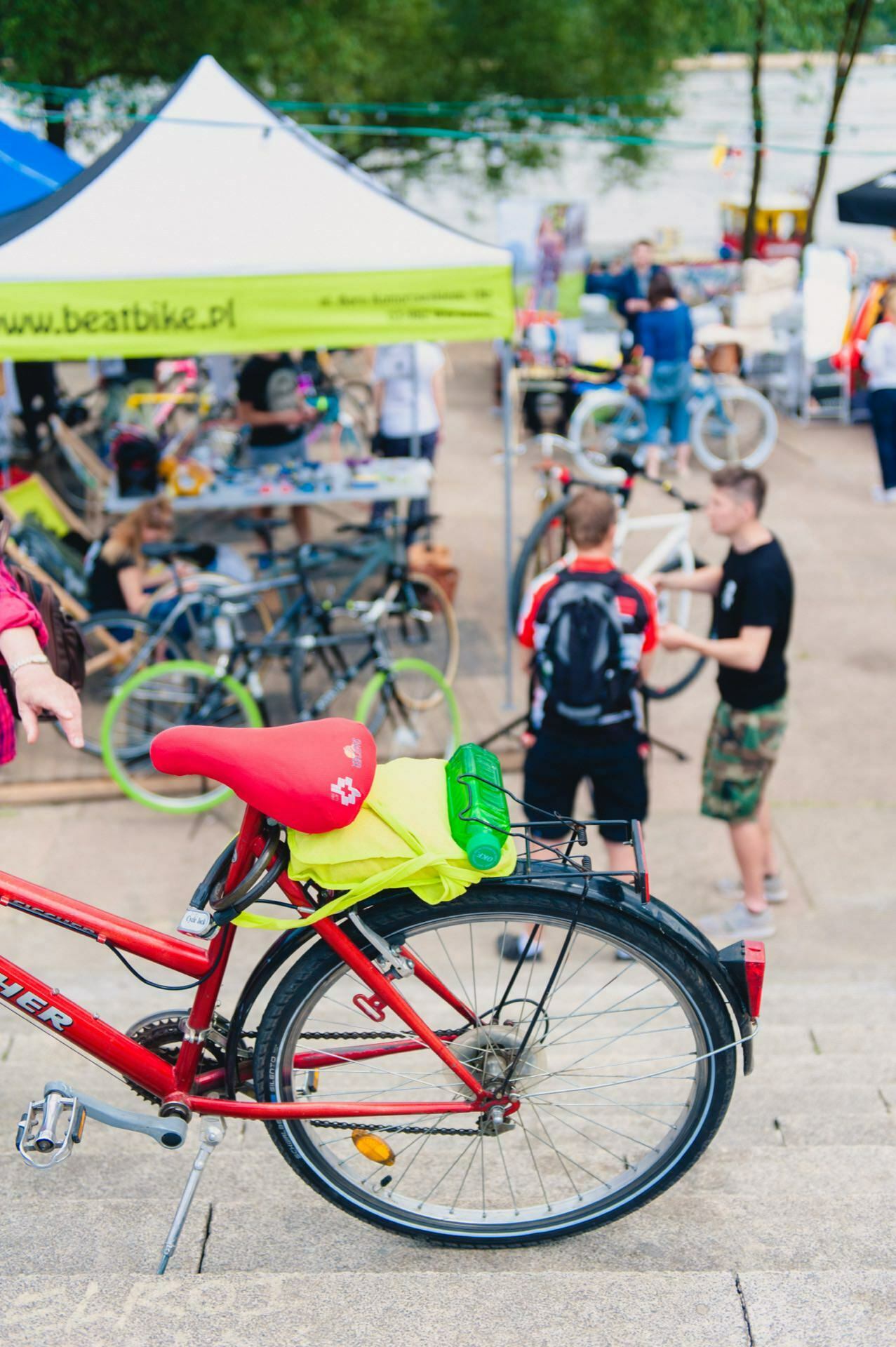 A red bicycle with a fluorescent green helmet and red saddle cap is parked on the steps. In the background is a vibrant outdoor event with people gathering around tents and bikes, perfect for a photo opportunity at the fair. The scene is bright and vibrant, capturing the essence of vibrant community gatherings.  