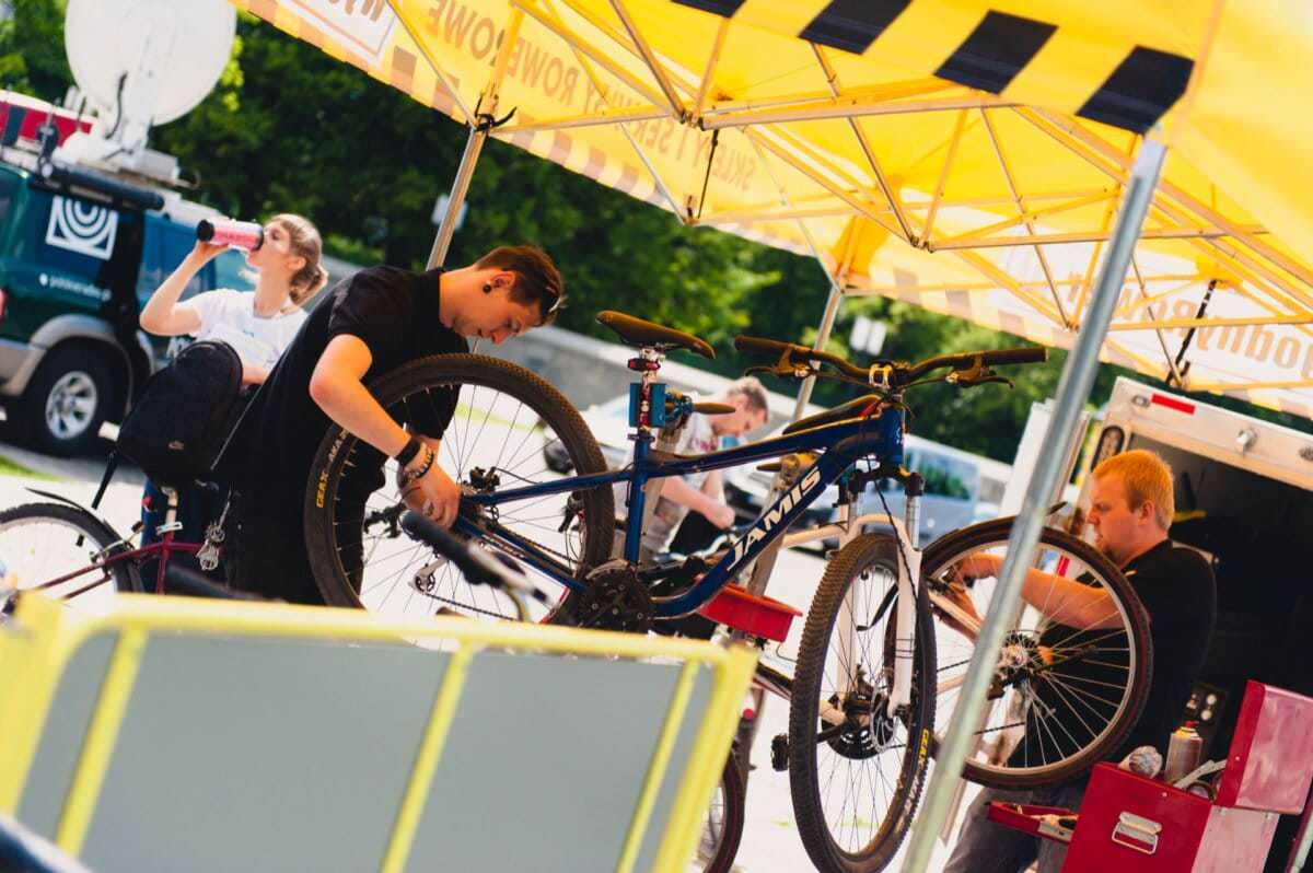 Dwóch mechaników rowerowych pracuje na rowerach pod żółtym namiotem z napisami „Specialized” i „Power” po bokach. Jeden mechanik reguluje przednie koło roweru górskiego, a drugi sprawdza koło innego roweru. W tej urzekającej fotorelacji z targów osoba w tle pije z butelki.