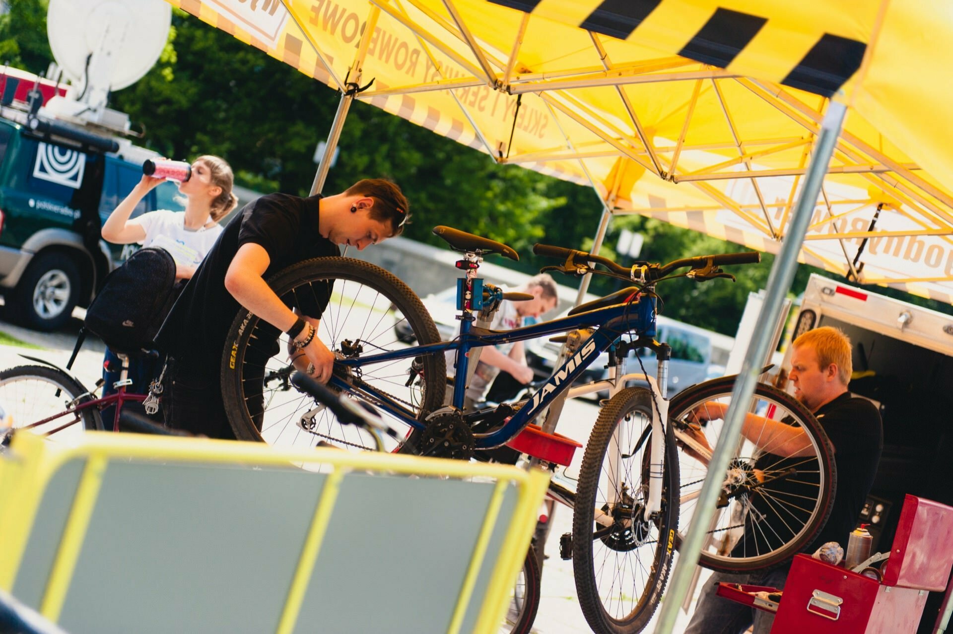 Two bicycle mechanics work on bicycles under a yellow tent with "Specialized" and "Power" signs on the sides. One mechanic adjusts the front wheel of a mountain bike, while the other checks the wheel of another bike. In this captivating photo essay of the fair, the person in the background is drinking from a bottle.  