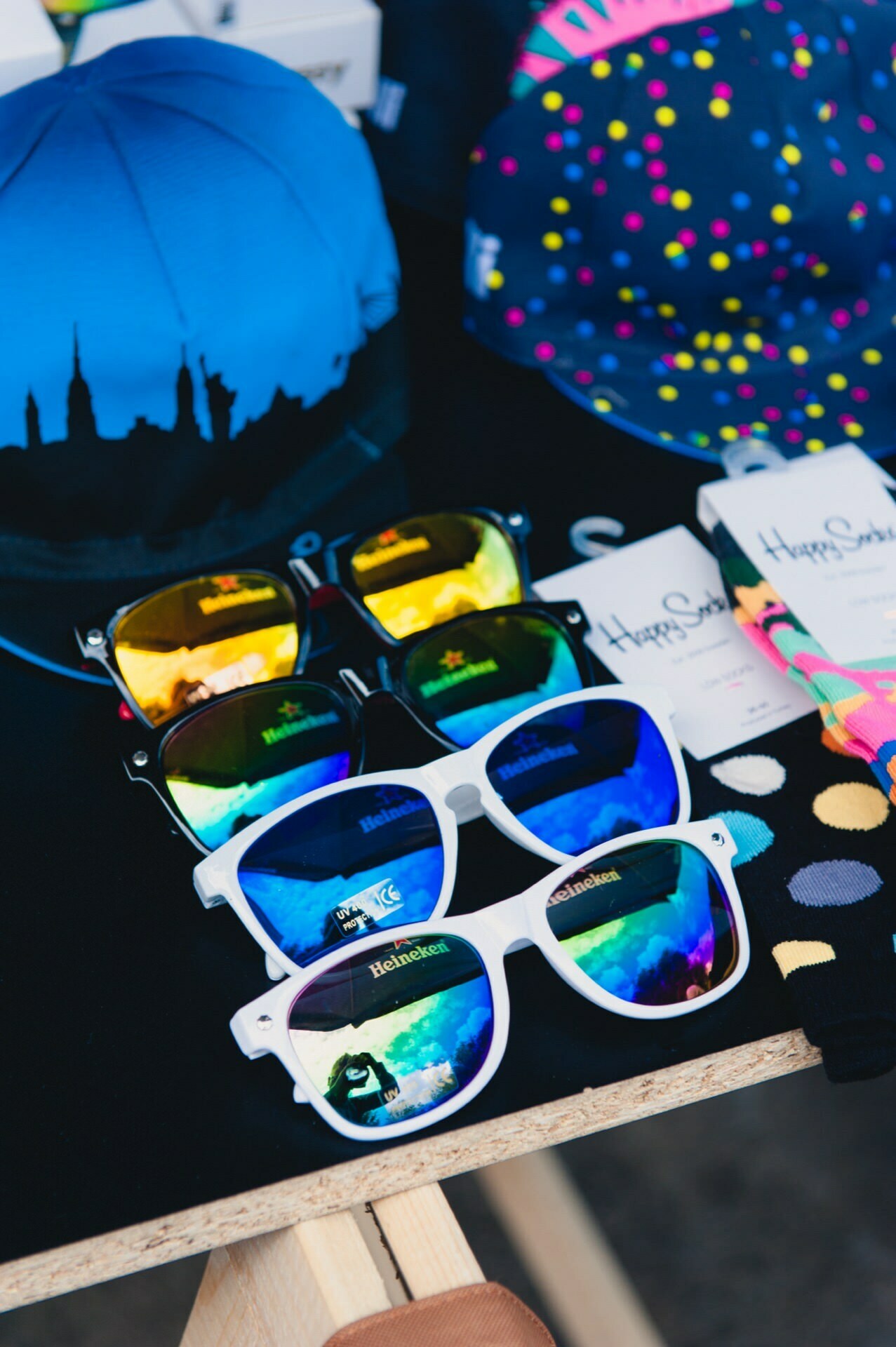 The vibrant photo report from the fair shows a display of colorful sunglasses on a table, consisting of three pairs with reflective lenses in black, white and yellow frames. Behind them are vibrant hats and multi-patterned Happy Socks in wrappers. The background is out of focus, typical of an event photographer.  