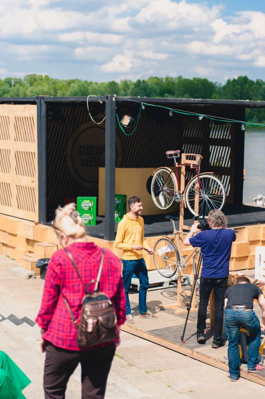 Grupa ludzi gromadzi się wokół drewnianego straganu nad brzegiem rzeki. Na stojakach znajdujących się wewnątrz stoiska wystawione są dwa rowery. Jedna osoba w żółtym swetrze rozmawia z innymi, druga w czerwonej kratce idzie w stronę straganu. Drzewa i częściowo zachmurzone niebo stanowią idealne tło dla tej fotorelacji z targów.
