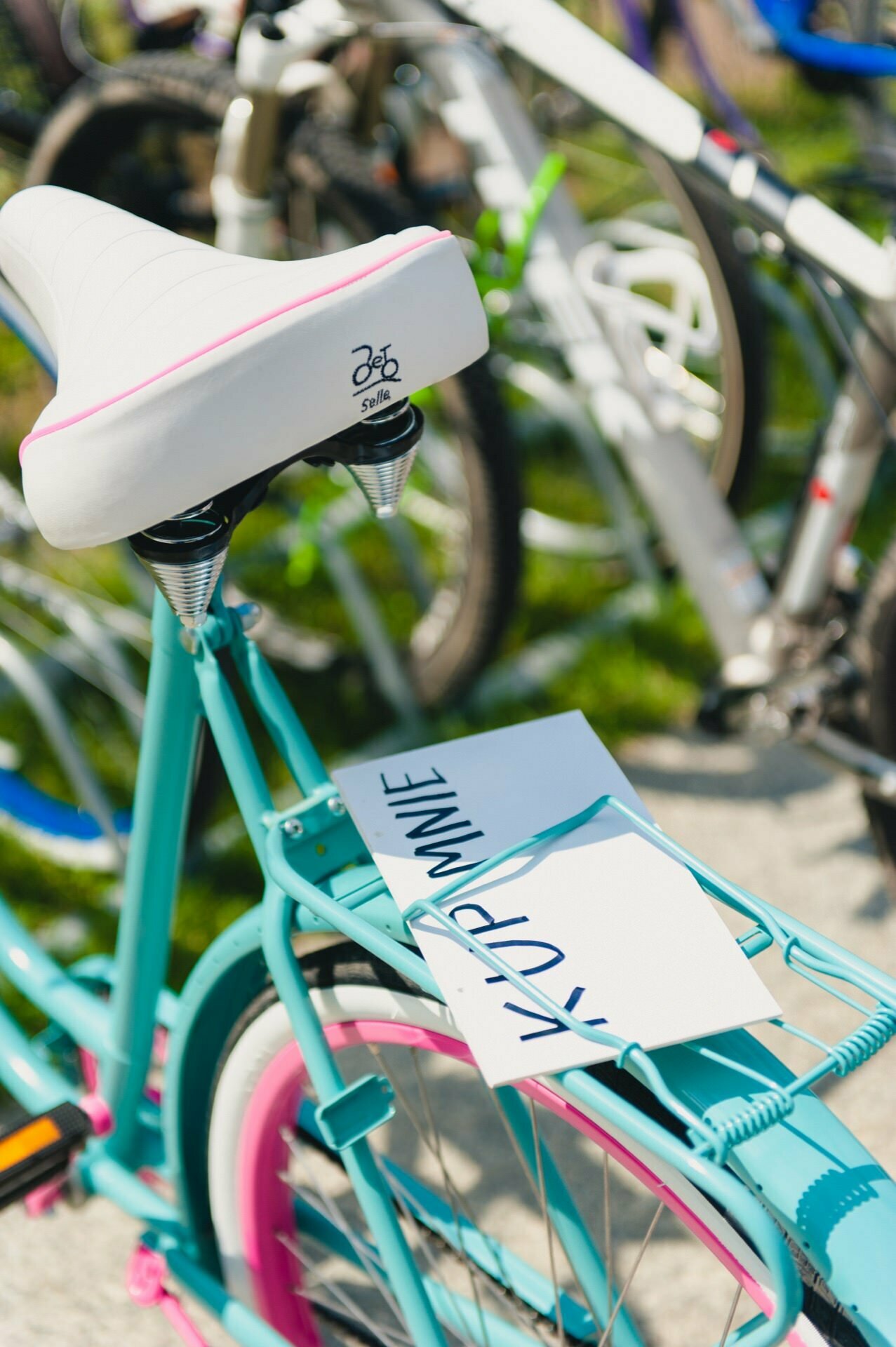 The turquoise bike with white and pink accents stands in a row with other bikes. On the back of the bike is a placard that reads "KÜP MUE" in large letters. The bike, likely captured by an event photographer, is equipped with a white saddle and rear rack.  