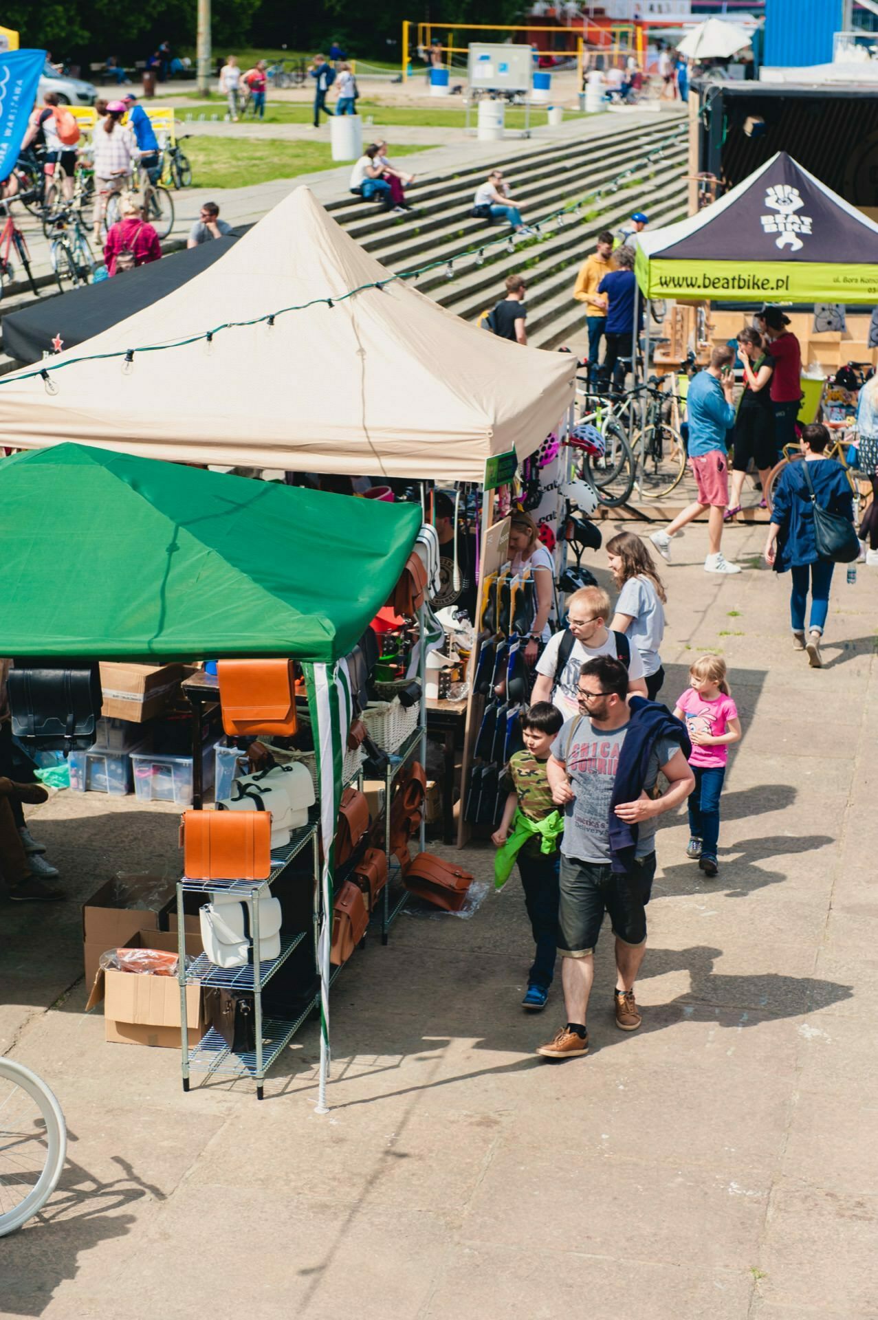 An outdoor market with several stalls selling various items. People, including families with children, walk around and shop, and an event photographer captures these moments. Some stalls have green or brown canopies, and bicycles can be seen in the background. The atmosphere is busy and lively.   