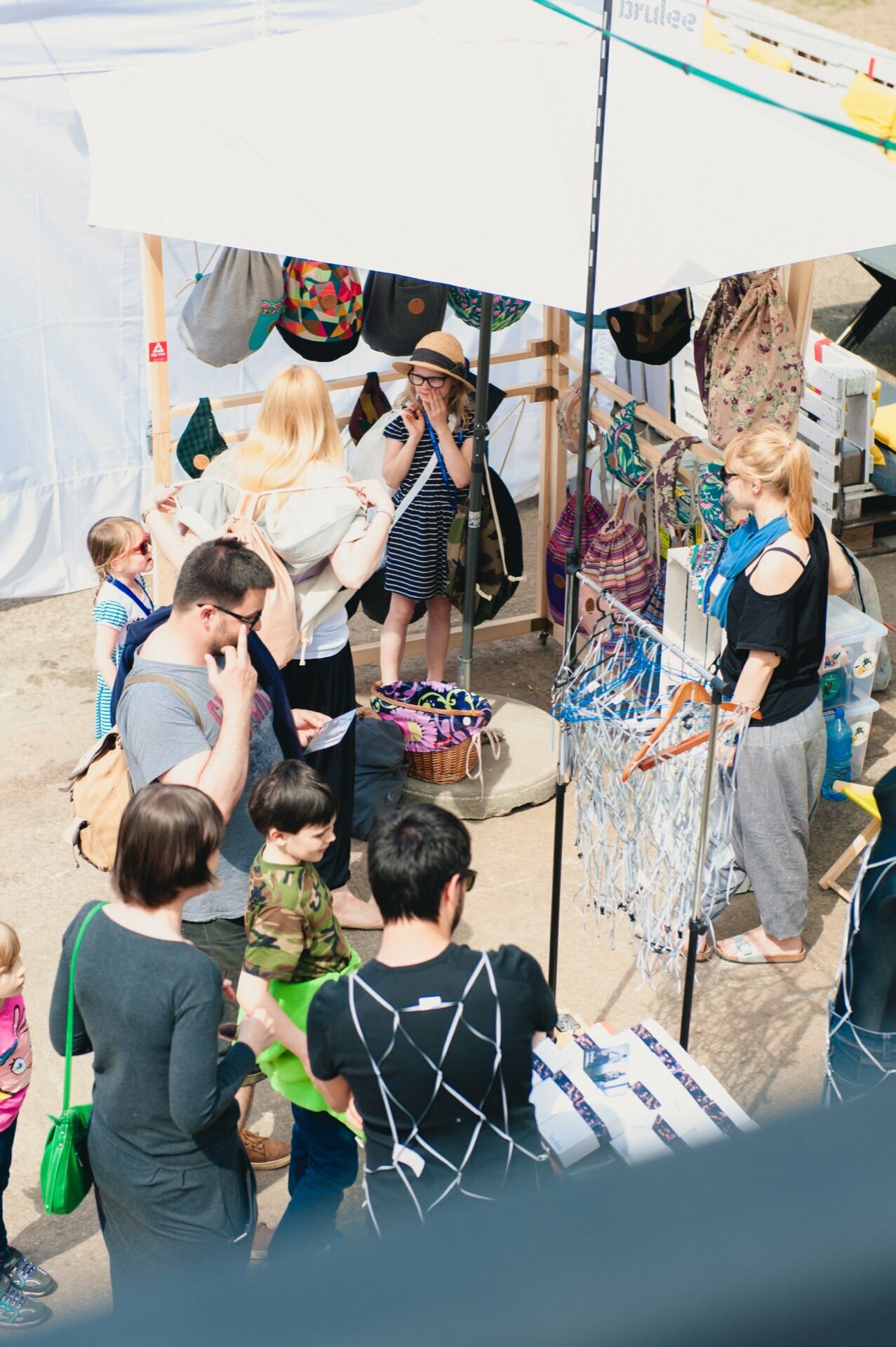 An outdoor market scene with people shopping at various stalls. A woman in the center serves customers, selling colorful bags and accessories under a white canopy. Families with children and other customers browse the stalls. The weather promises to be sunny and bright, perfect for a photo opportunity at the fair.   