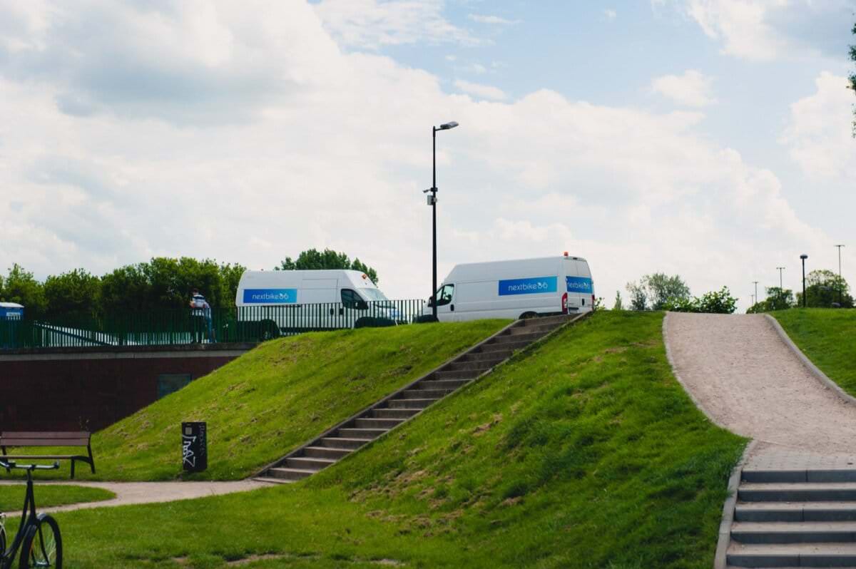 Dwie białe furgonetki z napisem „NHS Test & Trace” na boku stoją zaparkowane na szczycie trawiastego wzgórza obok brukowanej ścieżki i schodów. Na pierwszym planie widoczna osoba i rower, utrwalające moment przypominający sesję zdjęciową eventową pod częściowo zachmurzonym niebem.
