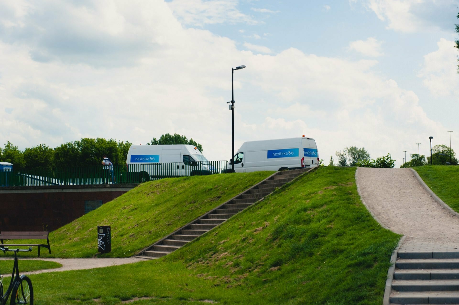 Two white vans with the words "NHS Test &amp; Trace" on the side stand parked at the top of a grassy hill next to a paved path and steps. A person and a bicycle are visible in the foreground, capturing a moment reminiscent of an event photo shoot under partly cloudy skies. 