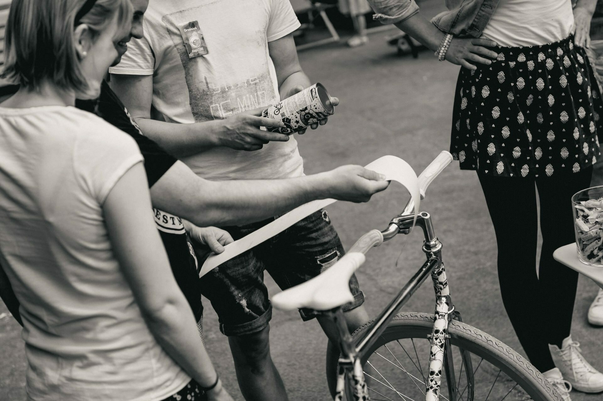 A group of people are gathered around a bicycle, with one person holding a roll of tape and another sticking it to the bike's handlebars. The scene appears to be taking place outdoors and everyone is casually dressed. The black-and-white photo captures the perfect moment that any event photographer would include in their photo story of the fair.  