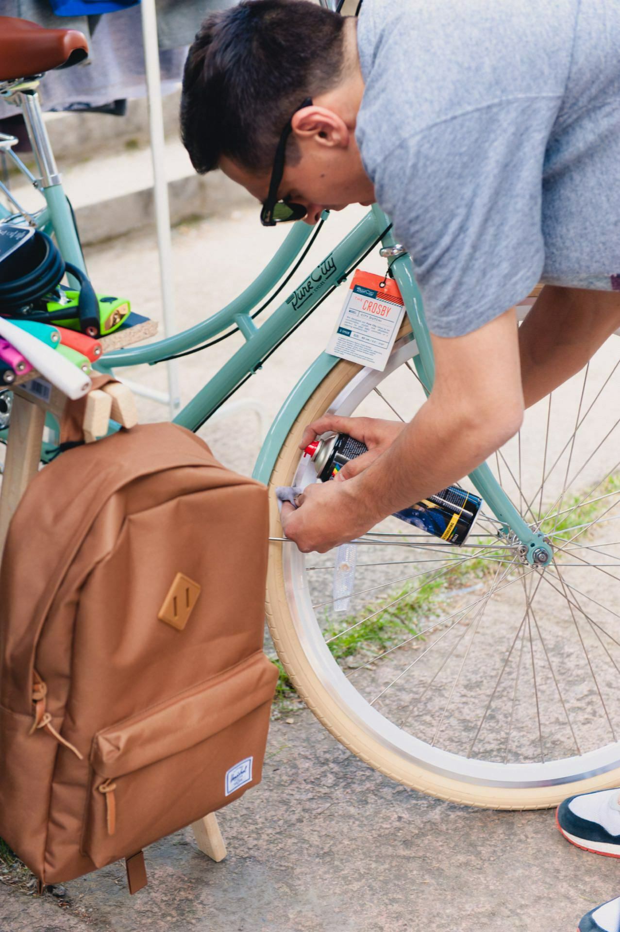 A person wearing a gray t-shirt and sunglasses adjusts a light blue bicycle with brown tires, parked on a stone surface. A brown backpack hangs next to the bike. The outdoor scene can be part of a photo shoot of a fair or captured by an event photographer in a bustling market or park.  