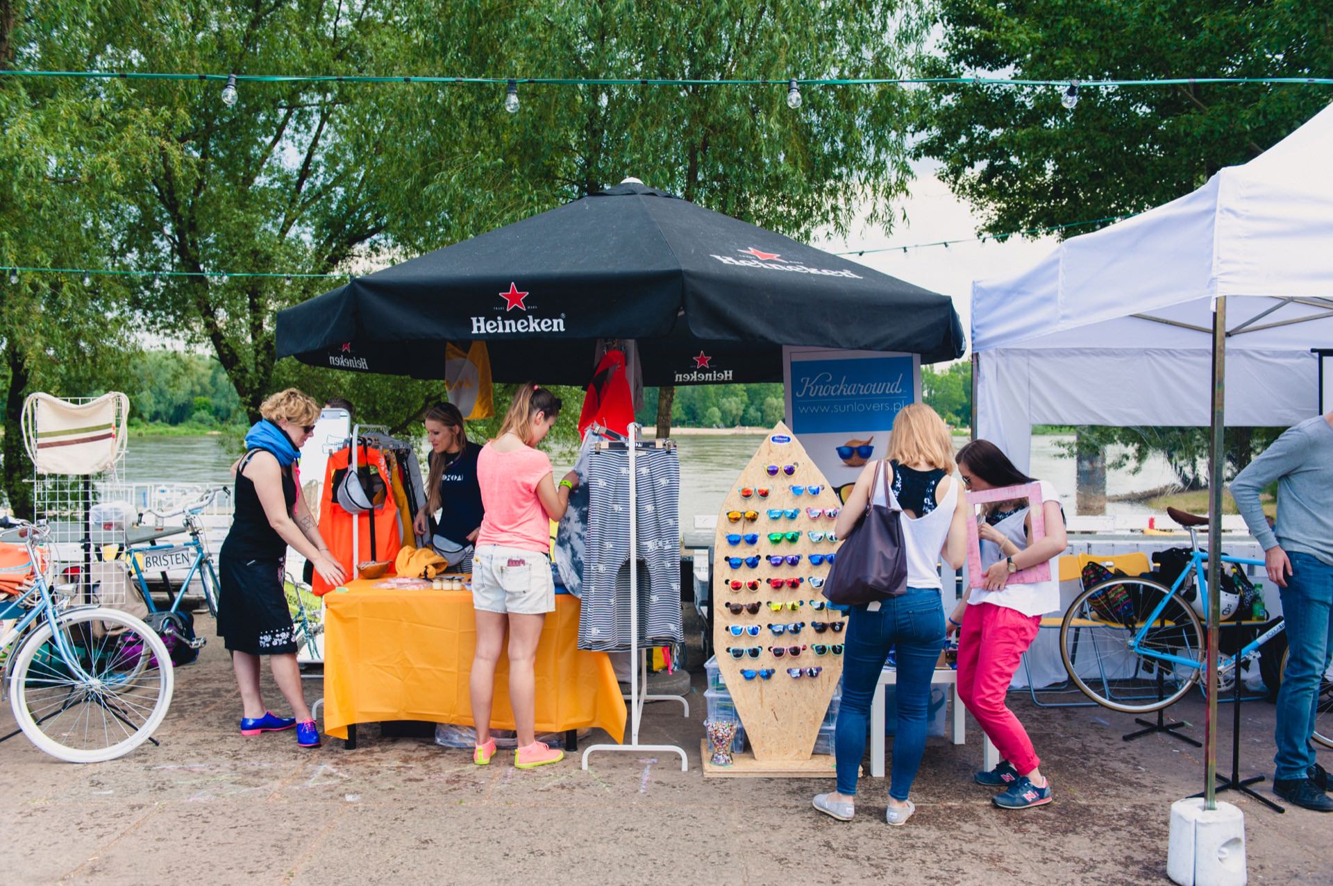 A lively outdoor booth under the Heineken brand umbrella, with various items for sale, including clothing, accessories and sunglasses. Several people are browsing the items, and bicycles are parked nearby. In the background, you can see trees and a large body of water - perfect for a photo opportunity at the fair.  