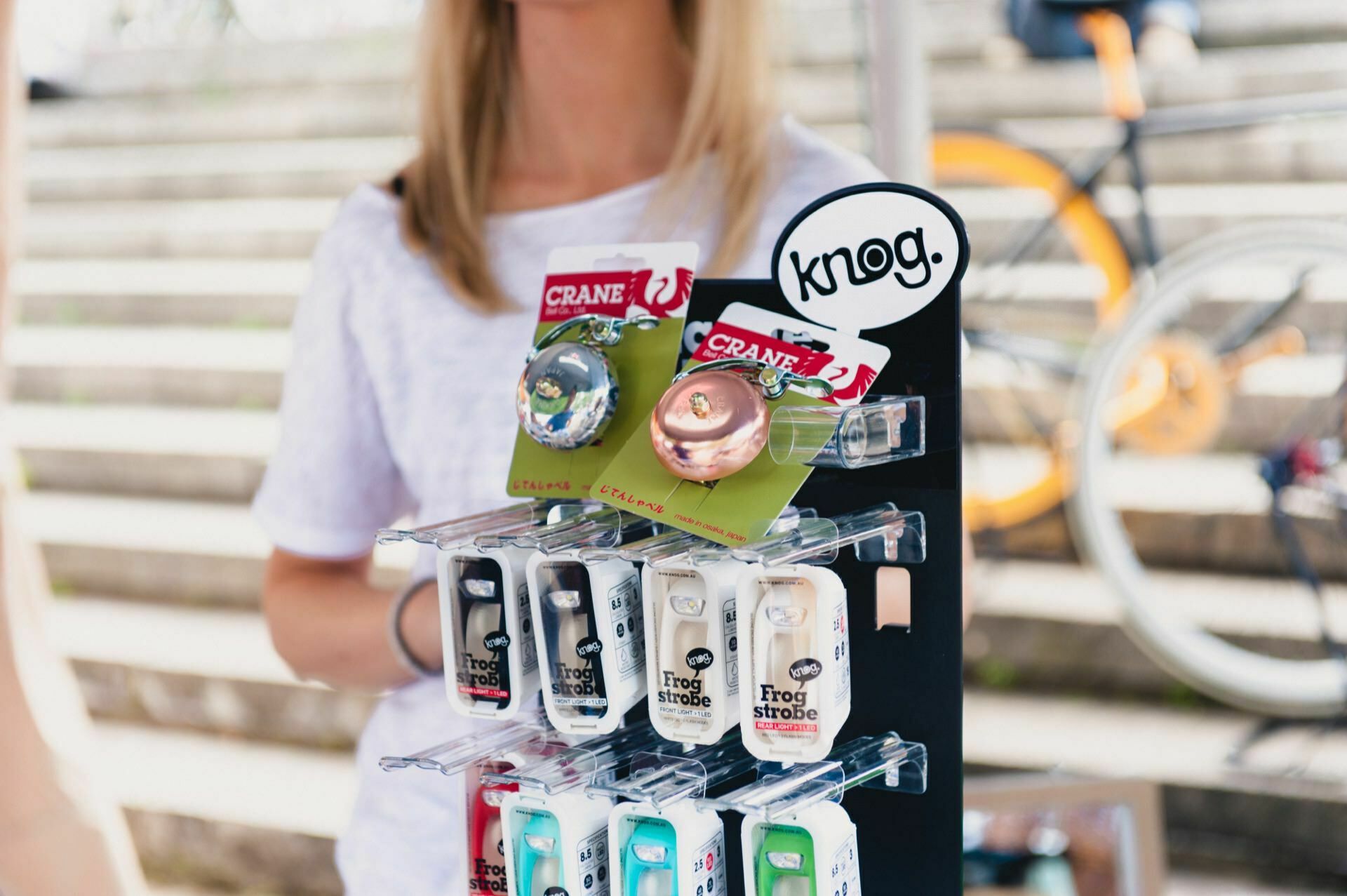 A close-up of an exhibition stand with various bicycle accessories, including bells and Frog Strobe lights from brands such as Crane and Knog. The stand, placed outdoors with a blurry background of a person and a bicycle, captures the essence of the show's photo coverage. 