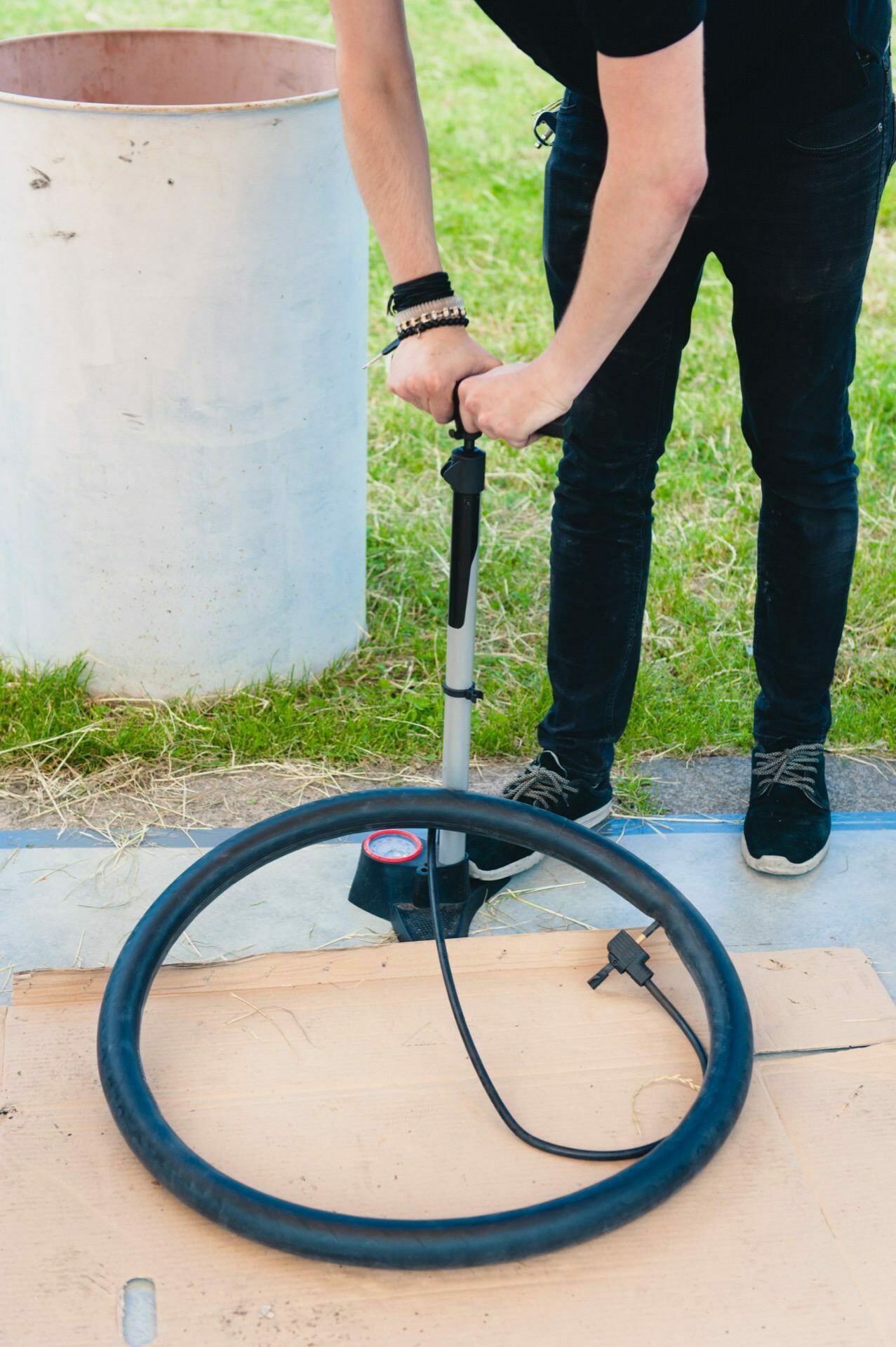 A person wearing a black shirt and wristbands is using a floor pump to inflate the inner tube in a bicycle tire. The scene, reminiscent of an event photographer capturing details of everyday life, takes place outdoors, with grass in the background and a white cylindrical object nearby. The inner tube rests on a cardboard box on the ground.  