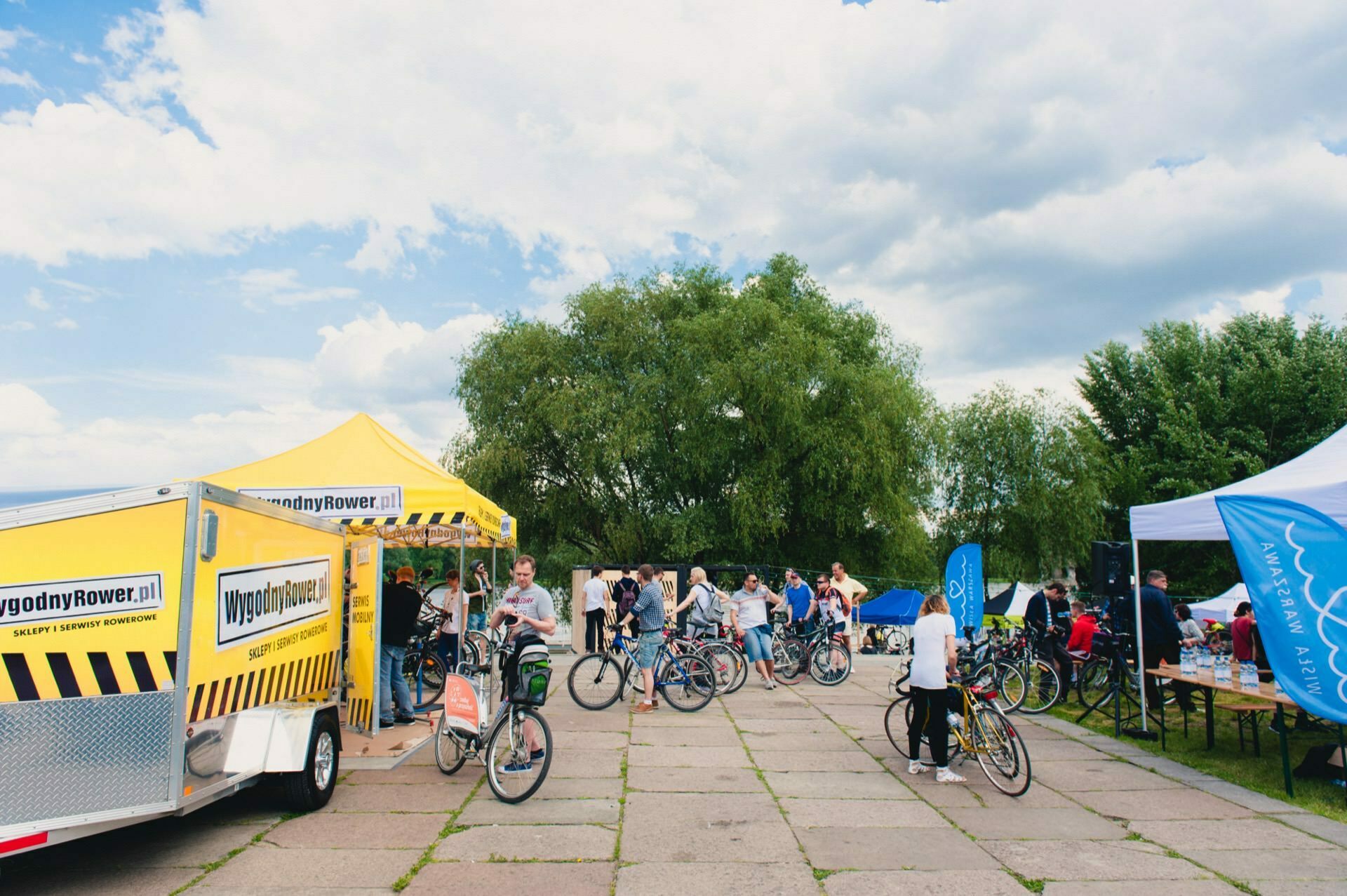 The lively outdoor event is a crowd of people gathered under a yellow tent and a truck with the sign "ConvenientRower.co.uk". Many participants are riding or looking at bicycles. The surrounding greenery and cloudy skies hint at a park-like setting, perfectly captured in the photo report of the fair on this pleasant day.  