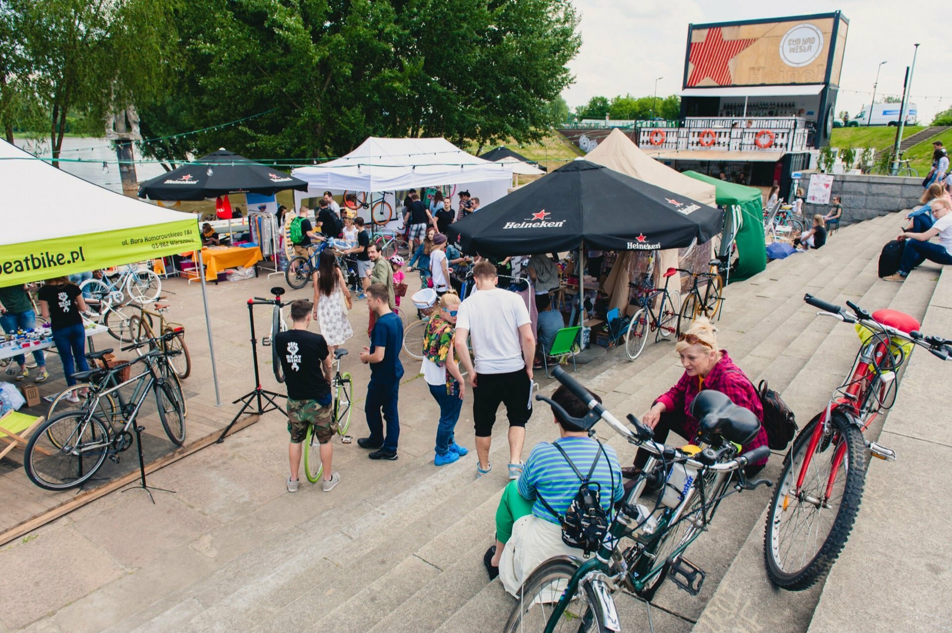 A lively outdoor cycling event with people browsing the various booths and tents, perfect for a photo-op of the fair. Several bicycles are parked or manned by individuals. In the background you can see trees and a multi-story structure as people stand, talk and socialize in the stairwell.  