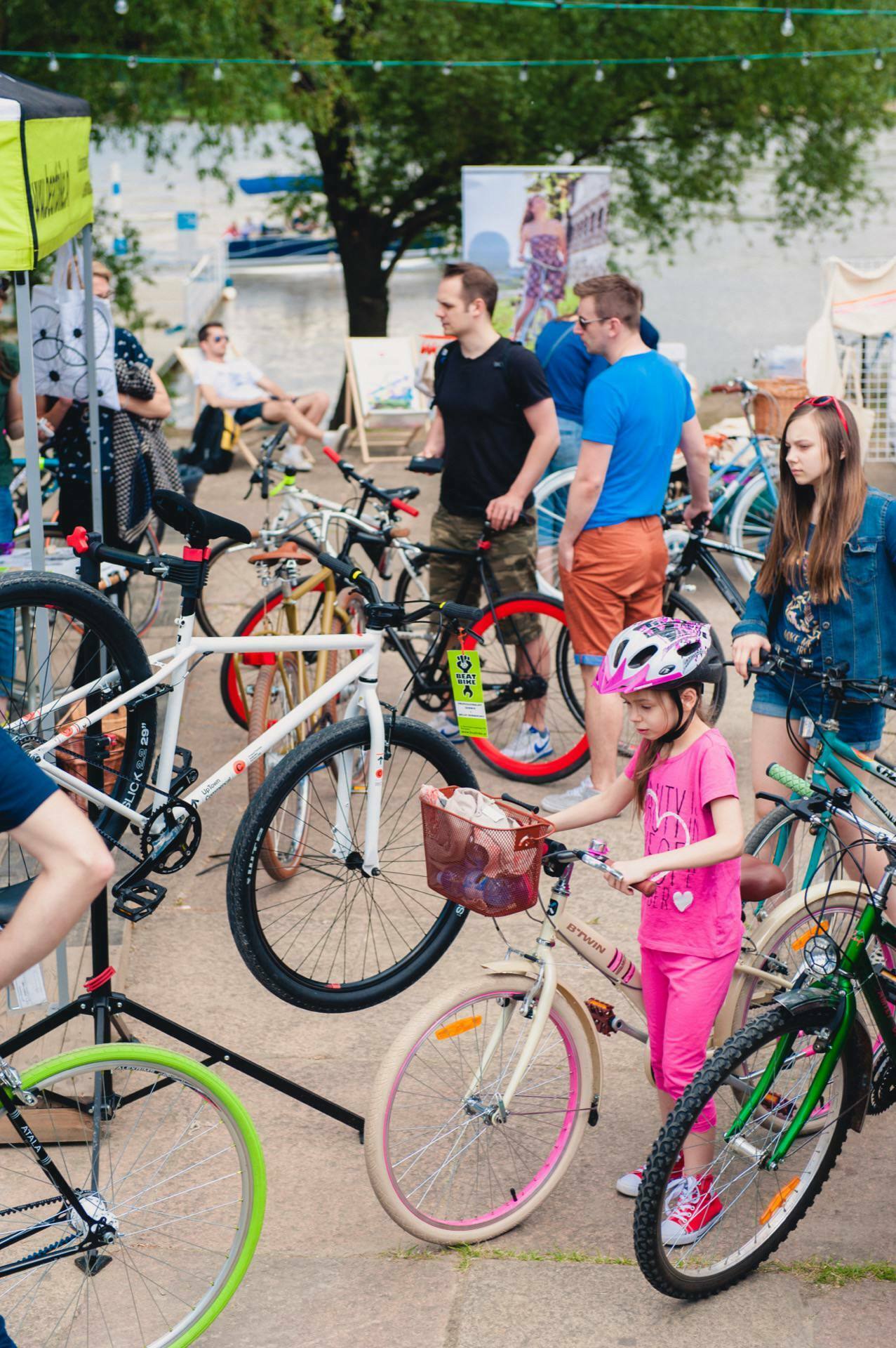 A group of people gather outside at a bike fair. Adults and children are looking at various bicycles displayed on racks. A young girl wearing a helmet and pink outfit stands next to a bicycle. In the background you can see the trees and the river, beautifully captured by our event photographer for this photo essay of the fair.   