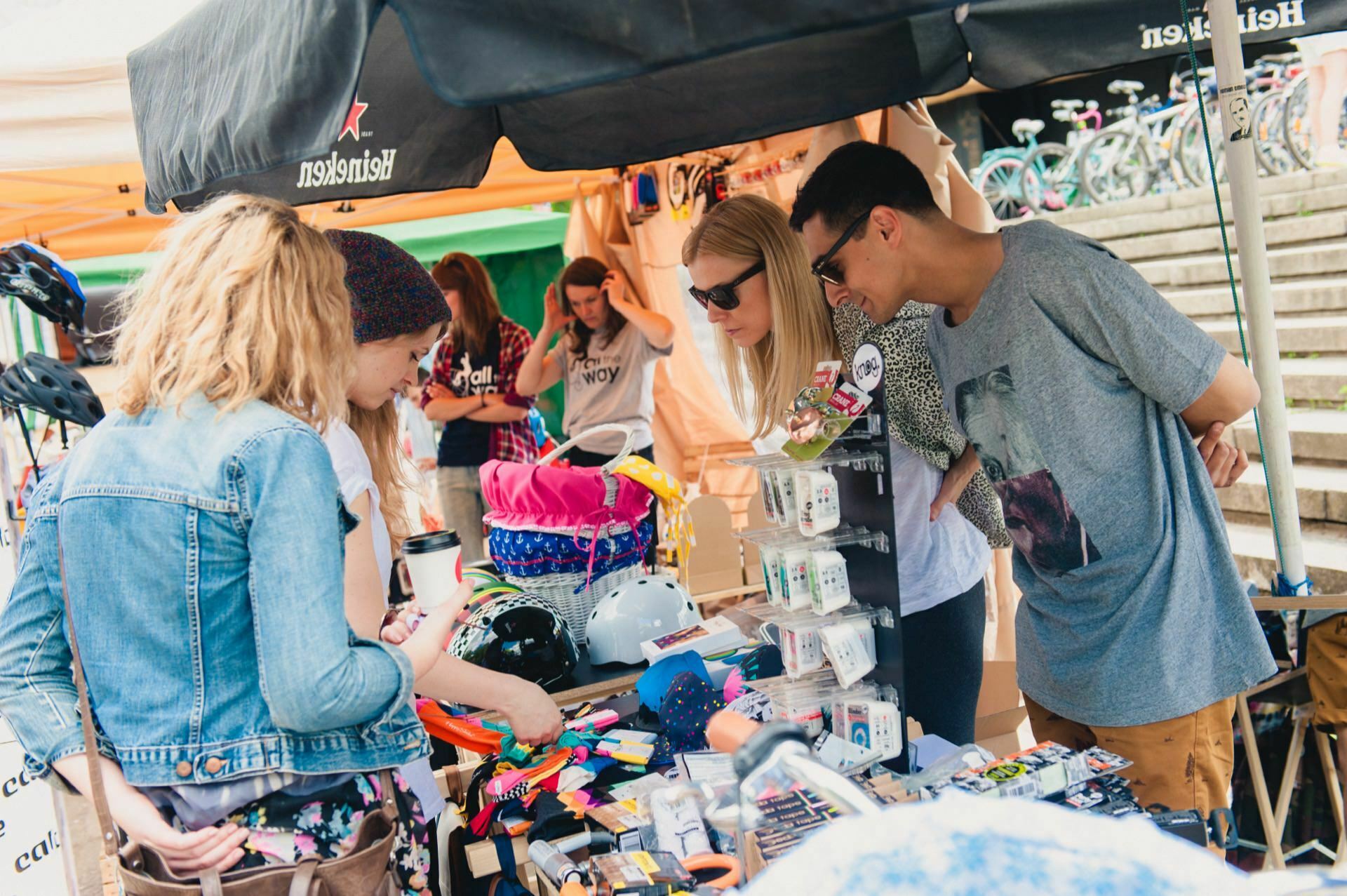 A group of people are browsing various colorful items on a table under a canopy at an outdoor market. Helmets, stickers and other small novelties are displayed on the table, and bicycles are visible in the background. The atmosphere seems lively and casual, perfect for a photo essay of the fair.  